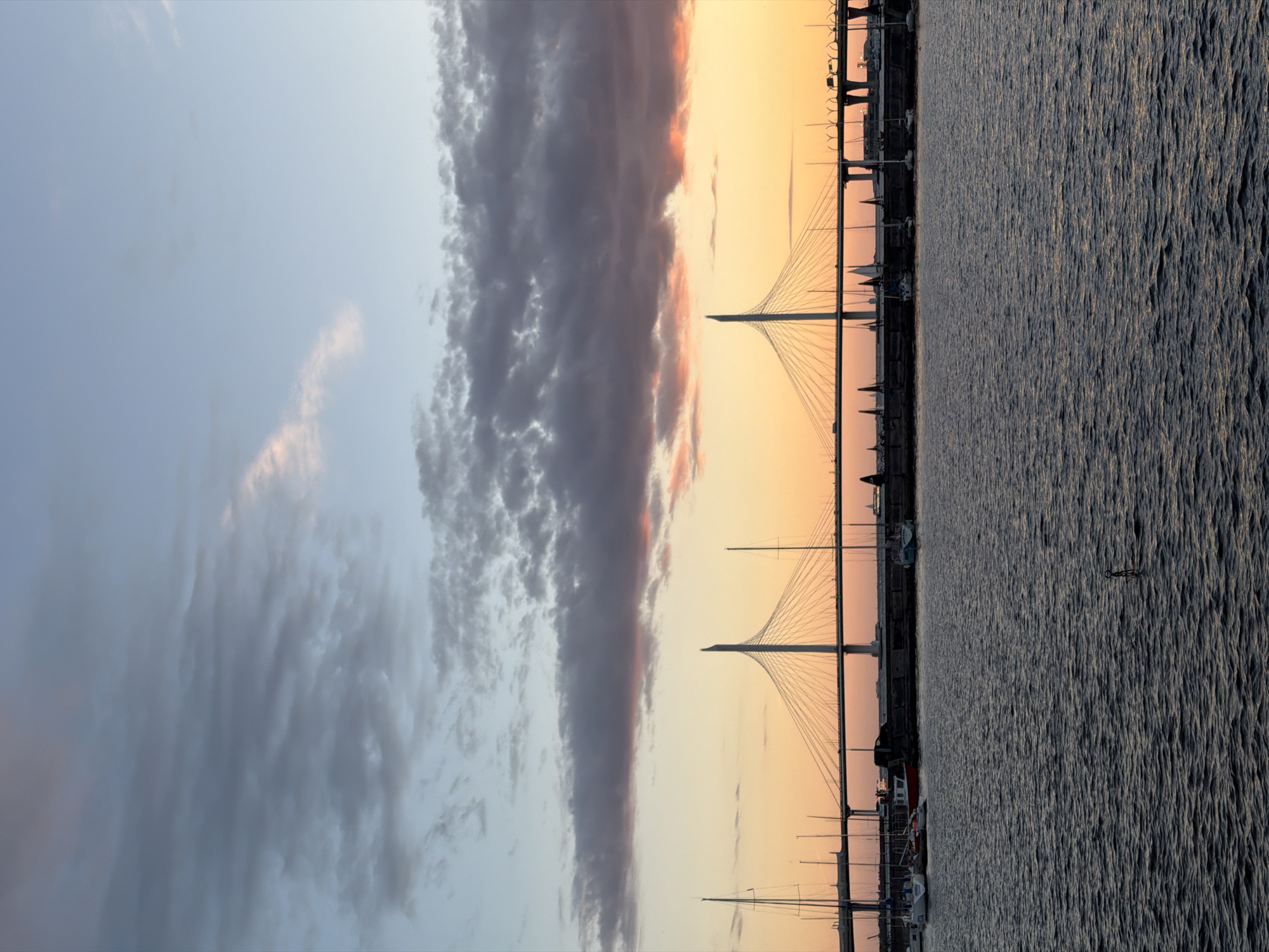 The image shows a cable-stayed bridge, likely the Western High-Speed Diameter in Saint Petersburg, Russia, at sunset. The sky is filled with a gradient of colors, from a pale blue at the top to soft oranges and pinks near the horizon. Dark, textured clouds are present in the upper portion of the sky. The bridge's distinctive pylons and cables are prominent against the colorful sky. Below the bridge, there is a body of water with several sailboats moored along the shore. The water is rippled, reflecting the colors of the sunset. There are also some vehicles visible on the bridge, indicating it is in use. The overall scene is one of a picturesque urban landscape at dusk.
