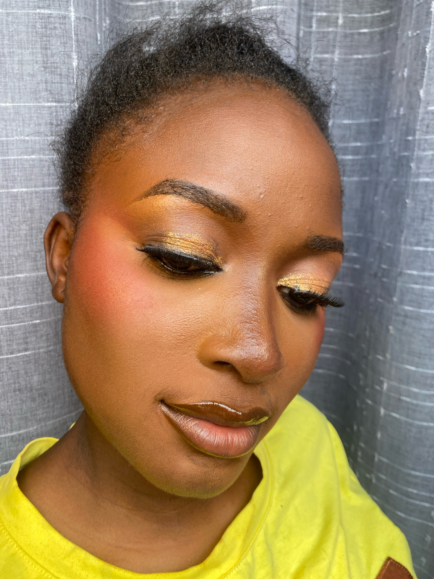 A close-up, medium shot captures a young Black woman from the chest up, her face angled downwards. She is wearing a bright yellow t-shirt and her dark, textured hair is pulled back. The background consists of a grey, pleated curtain. Her makeup features prominent orange blush on her cheeks, gold glitter eyeshadow, winged eyeliner, and voluminous false eyelashes. Her lips are painted with a glossy, brown lipstick. The lighting is soft, highlighting the textures of her skin and makeup. The image appears to be taken indoors.