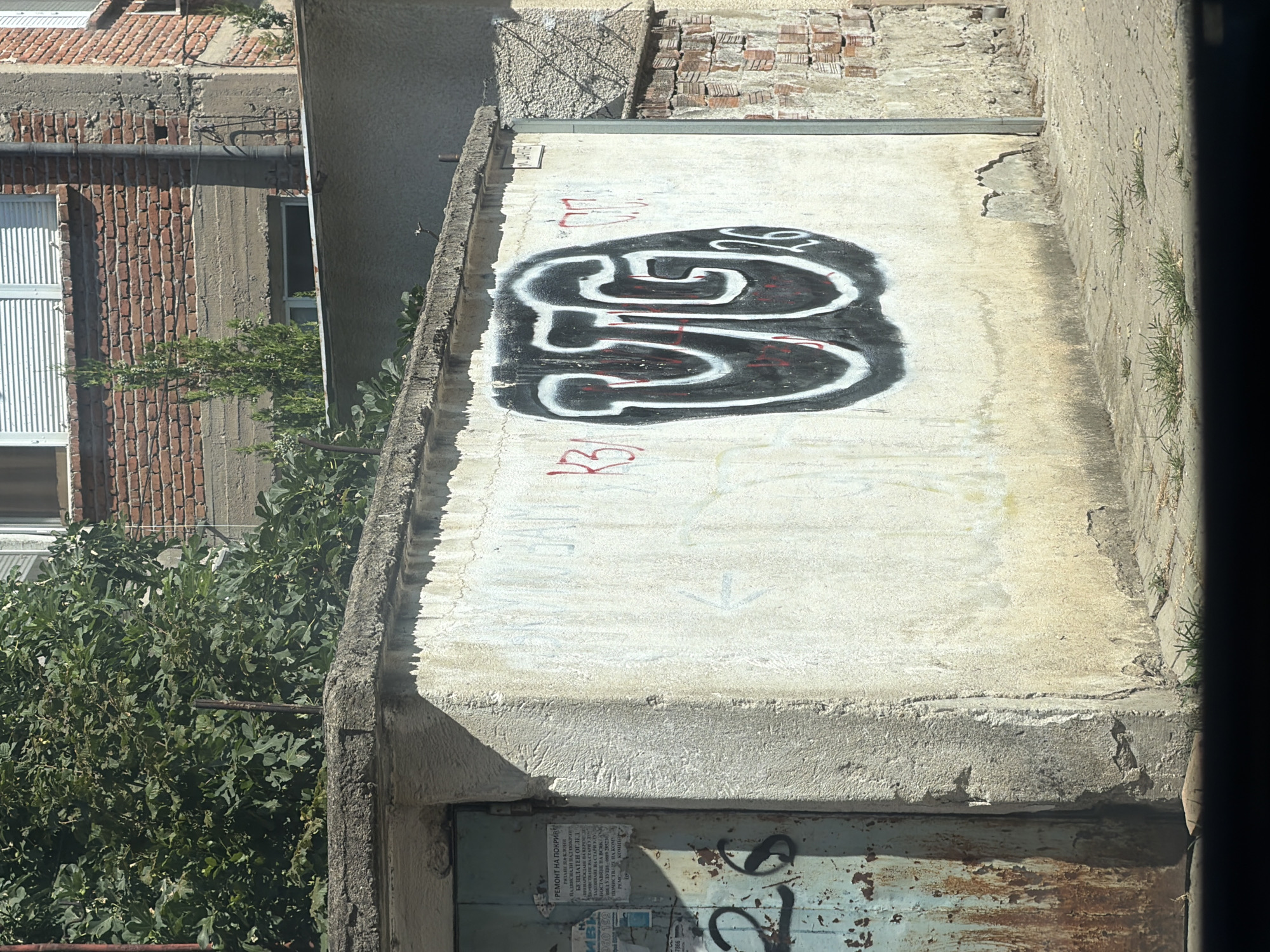 The image depicts an outdoor scene on a sunny day, likely in Plovdiv, Bulgaria, given the context. A concrete structure, possibly a garage or wall, dominates the foreground. The wall is covered in graffiti, most notably a large, bold black and white stylized "UTG" tag with red accents. To the left of this main tag, there are some red graffiti marks including "KB31" and what appears to be "017". Near the left edge of the wall, a green metal door is visible, heavily weathered and rusted, with some faded signage and the black graffiti numbers "26" spray-painted on it. The wall itself is a light, off-white color, with areas of staining and rough texture. Behind the concrete structure, a building constructed of red brick is visible, with several windows. Lush green foliage, likely from a tree, partially obscures the brick building and the top of the concrete structure. The ground in front of the wall is paved with interlocking bricks and has patches of grass growing through the cracks. The overall impression is one of urban decay and street art.