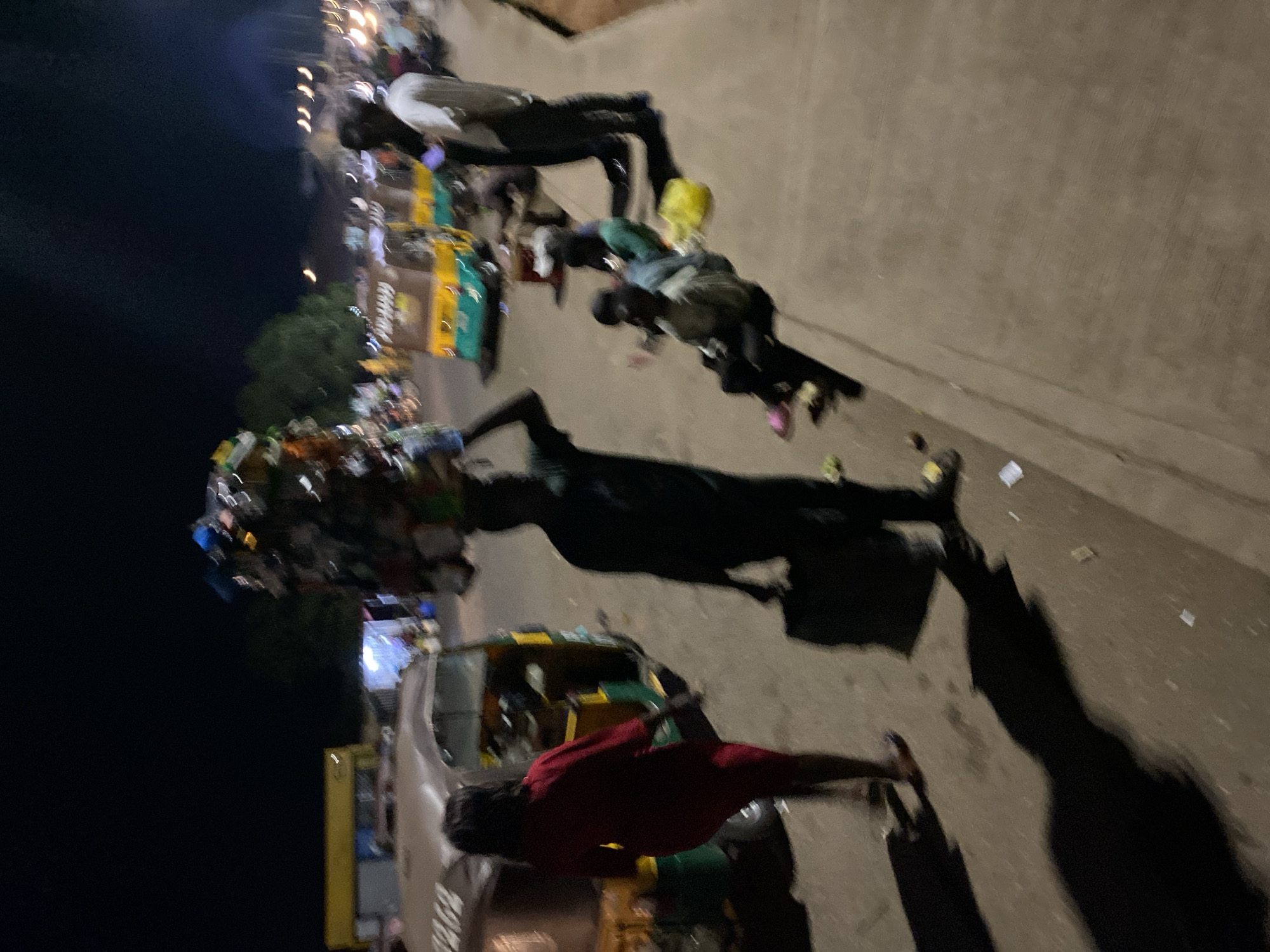 At night in Bukuru, Nigeria, the scene captures a bustling street. Several people are walking along a sidewalk. A man carries a large bundle on his head, casting a significant shadow. A red vehicle, potentially a taxi or auto-rickshaw, is visible, and other vehicles appear further down the street. The environment is illuminated by streetlights. The street is littered with small pieces of trash.