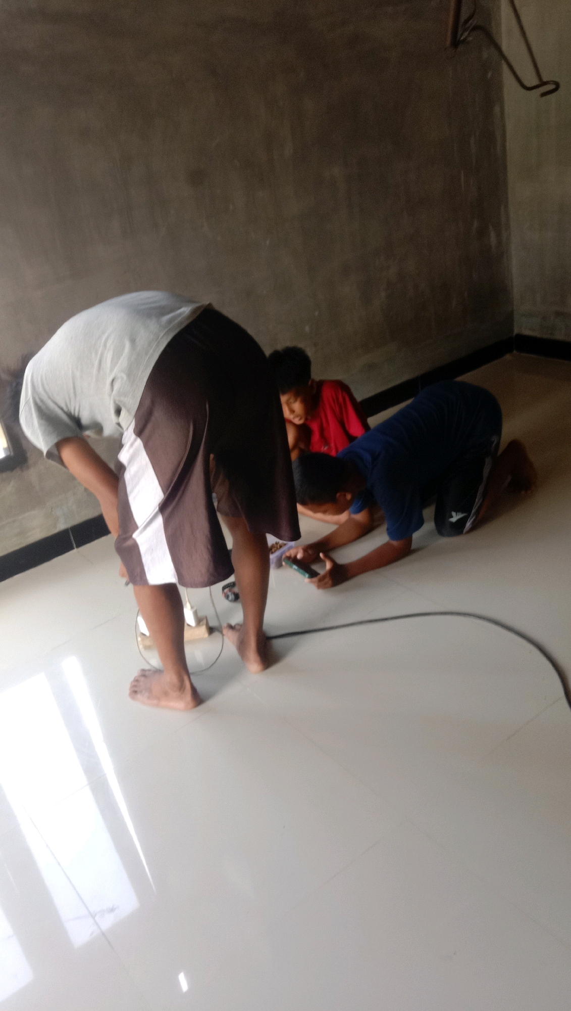 Three young males are on the floor of a room with a gray concrete wall and a shiny white tile floor. One male, wearing a gray t-shirt and brown shorts with white stripes, is bent over with his back to the camera, appearing to be working on something on the floor. Another male, wearing a red t-shirt, is seated on the floor behind the first male, looking towards the right. A third male, wearing a blue t-shirt and dark shorts, is kneeling on the floor in front of the first male and to the right, holding a mobile phone and looking at it. Wires are visible on the floor. The scene takes place indoors, likely in a residential setting in Kota Pari, Indonesia.