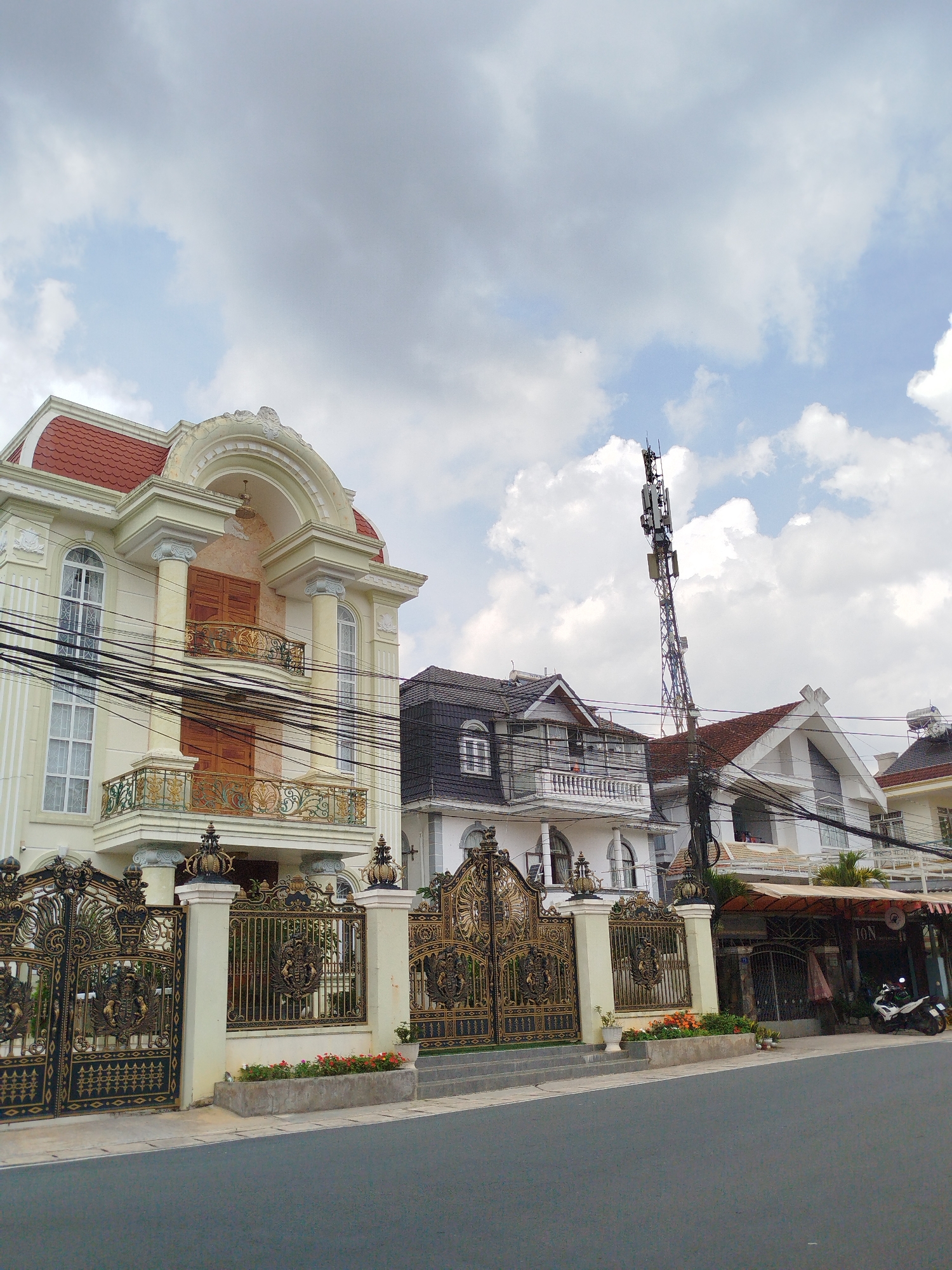 The image captures a street scene in Lâm Viên - Đà Lạt, Vietnam. In the foreground, a gray asphalt road is visible. To the left, a cream-colored, multi-story building with a red tiled roof is prominently featured. This building has ornate golden balconies and elaborate, dark metal gates adorned with crowns and crests. Adjacent to it, to the right, are other buildings with similar architectural styles, featuring dark tiled roofs and balconies. A tall telecommunications tower stands between the buildings, topped with antennas. Power lines are strung across the scene, crossing in front of the buildings. Further to the right, a motorcycle is parked near the entrance of a business with an awning overhead. The sky is partly cloudy, with a mix of gray and white clouds. The overall impression is of a well-maintained residential or commercial street in a city with a distinct architectural aesthetic.