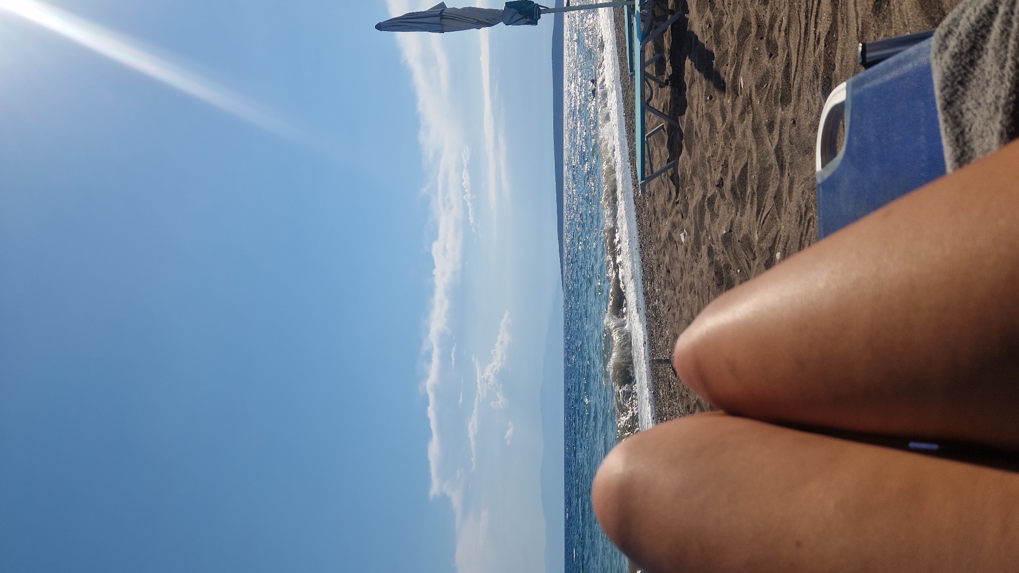The image depicts a view from a reclining position on a beach in the Municipal Unit of Asini, Greece, looking out towards the Aegean Sea. In the foreground, the tanned legs of a person are visible, resting on a blue lounge chair with a gray towel. The beach is composed of dark sand and pebbles. In the middle ground, the sea is a deep blue with gentle waves breaking on the shore. A row of empty, light blue sun loungers with a closed umbrella can be seen on the sand to the right. In the background, a hazy coastline with mountains is visible under a bright, sunny sky with scattered white clouds and a strong sunbeam emanating from the upper right corner. The sun is creating a glare on the surface of the water.