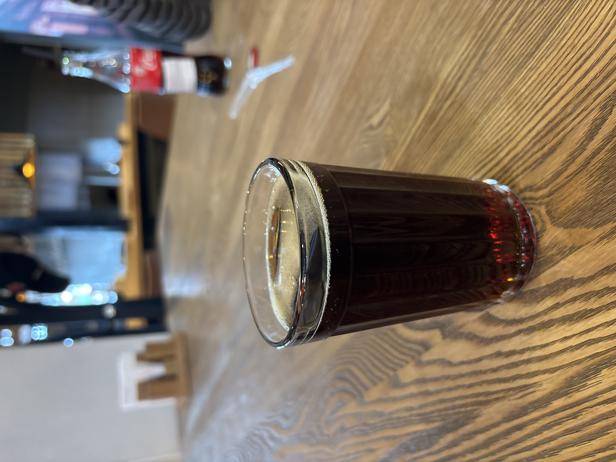 An overhead shot shows a glass filled with a dark carbonated beverage, such as cola, on a wooden table. The glass is partially obscured by the edge of the frame on the right. In the background, a bottle of Coca-Cola stands upright. The scene appears to be set in a cafe or restaurant. No people are visible in the image. The ambient lighting suggests an indoor environment, possibly with natural light filtering in from windows. The table's surface has a distinctive wood grain pattern. The location is identified as Zakharovo, Russia.