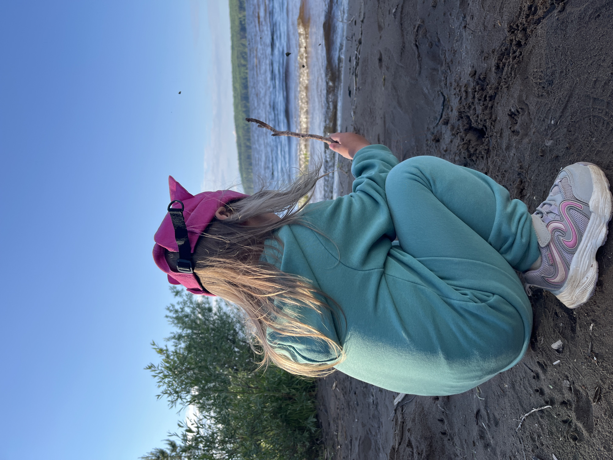 A young person, possibly a child, with long, light-colored hair is depicted crouching on a dark, wet sandy or muddy shore. They are wearing a bright pink cap with a black adjustable strap and a teal or mint green sweatsuit. Their right hand holds a thin wooden stick, which they are pointing towards the ground, suggesting an activity of drawing or playing in the sand at the water's edge. The person's feet are clad in grey athletic shoes with pink accents, and the word "combi" is visible on the side of one shoe.
The background features a calm body of water, likely a river or reservoir, with sunlight reflecting off its surface. A green, tree-lined bank is visible on the distant shore. The sky overhead is clear and a vibrant blue, with a couple of small, dark specks that may be insects or birds. To the left, some green foliage from a bush or tree can be seen. The scene is bright, indicating a sunny day, likely during the late morning or afternoon. The overall setting suggests an outdoor activity by the water in Chaikovsky, Russia.