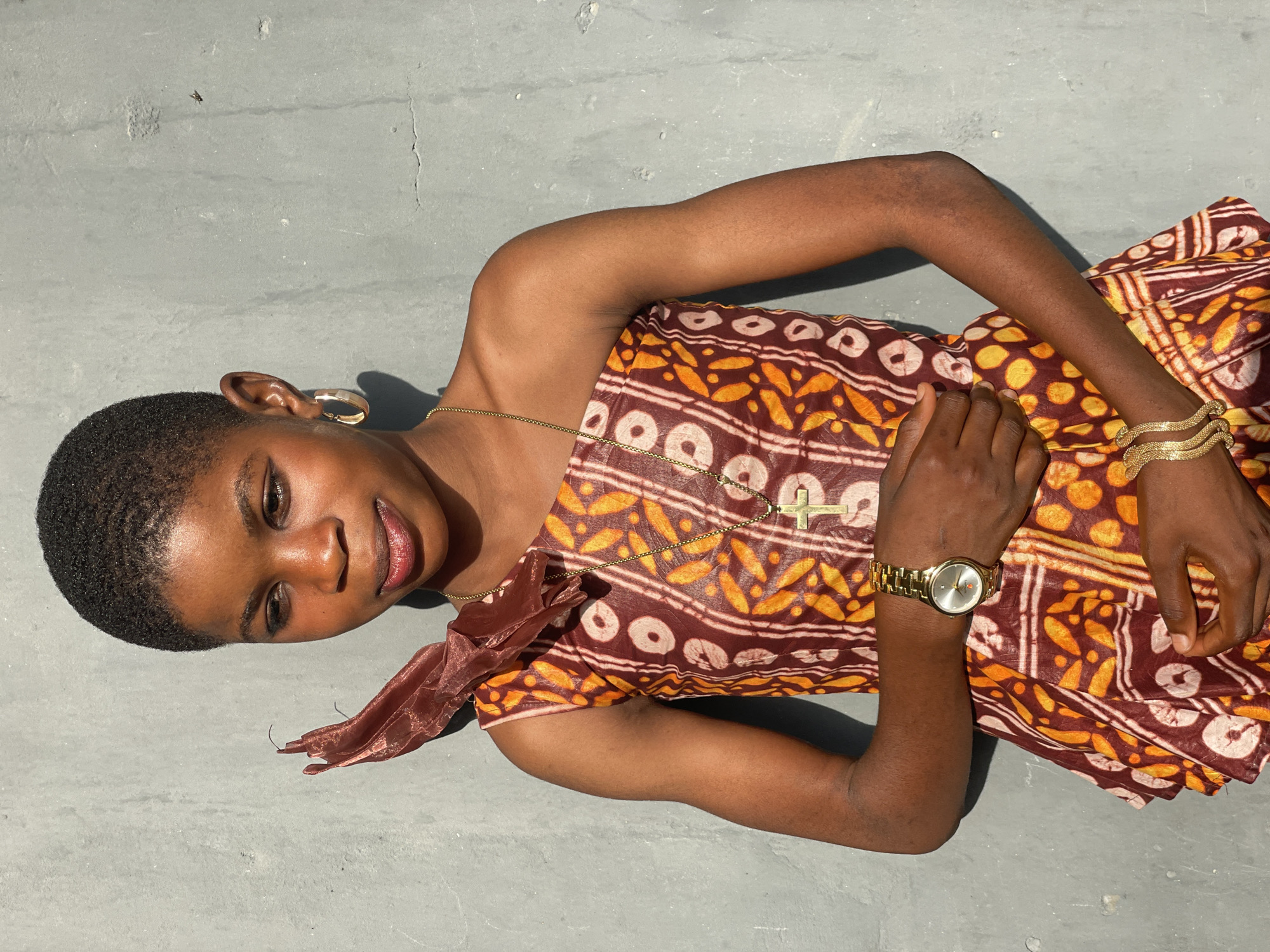 A young woman with dark skin and short, closely cropped hair is lying on a light gray, possibly concrete or tiled, surface. She is positioned diagonally across the frame, looking up and smiling subtly towards the viewer.
She wears a patterned dress in warm earthy tones of maroon/brown and orange/yellow, featuring circular and abstract organic shapes, suggestive of traditional African textile designs. The dress appears to be an off-the-shoulder style on one side.
Her accessories include gold-colored hoop earrings, a delicate gold chain necklace with a cross pendant resting on her chest, a gold-toned watch on her left wrist, and multiple stacked gold-toned bangles on her right wrist. Her makeup is subtle, with a reddish tint on her lips and some eye makeup.
Her right arm is bent, with her hand resting gently on the dress near her chest, while her left arm is also bent at the elbow.
The setting appears to be outdoors, with strong, direct sunlight creating sharp shadows and highlights on her skin and the ground, indicating a bright day, possibly midday or early afternoon. The ground surface is textured, showing minor cracks and imperfections. A small fly is visible on the ground above her head in some detail views. No visible text is present in the image. The overall mood conveyed is calm and pleasant.