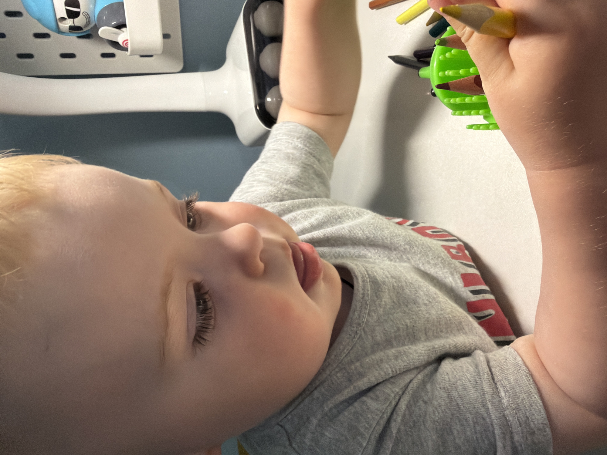 A young child is shown close-up, holding a green pencil holder with colored pencils. The child is fair-skinned with blond hair and long eyelashes. They are wearing a grey t-shirt with red and black stripes on the sleeve. In the background, there is a white wall with a blue section and some blurred white objects, including what appears to be a container with two small cartoon figures. The scene is likely indoors, but the location in Votkinsk, Russia, cannot be determined from the image.
