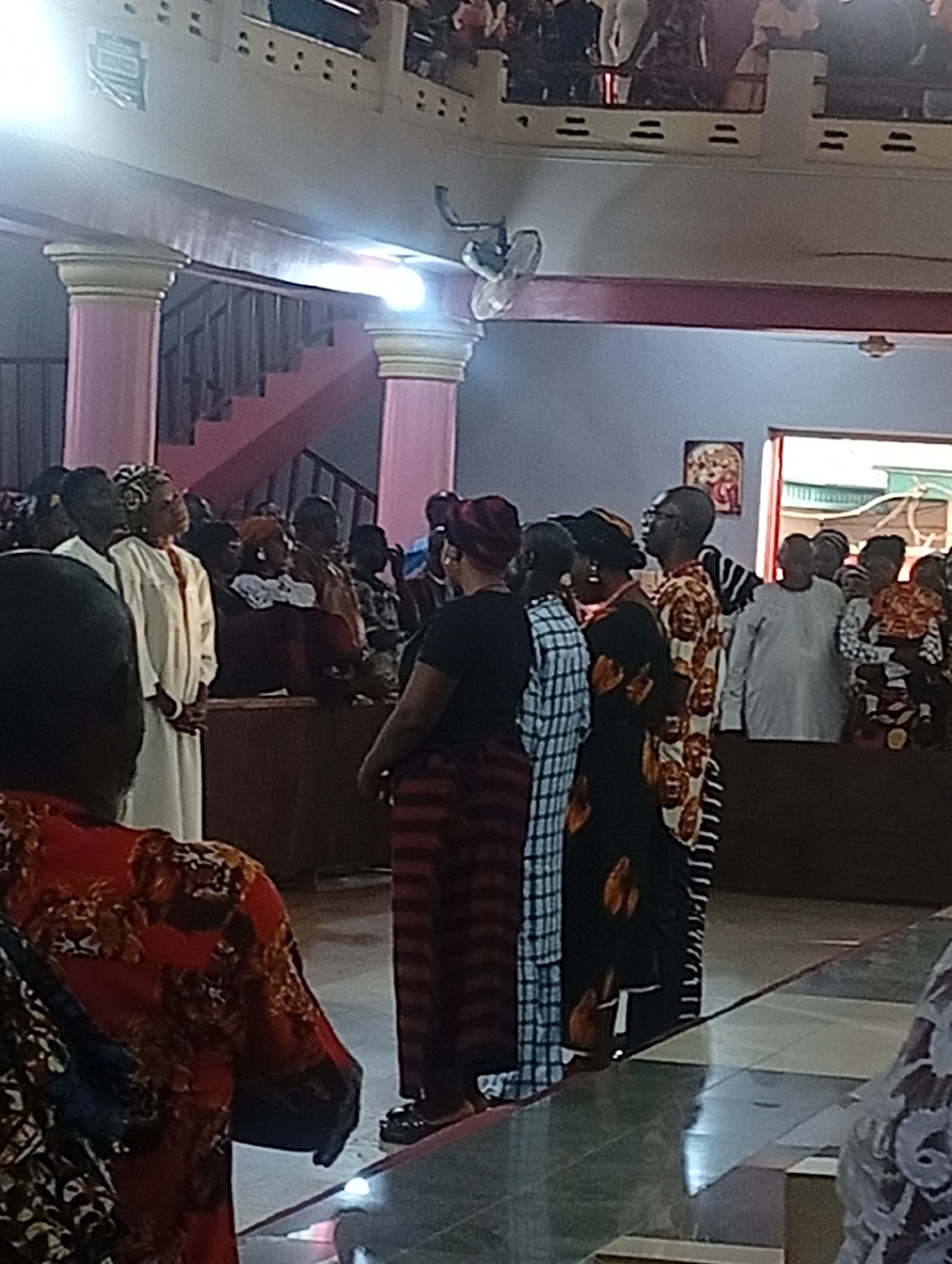 The image shows a crowded indoor scene, possibly a church or community hall in Unknown, Unknown. A group of people are gathered, with several individuals standing in the foreground. Their attire varies, including patterned dresses, white robes, and colorful headwraps. The setting includes architectural details such as pink columns, stairs, and a balcony with people watching from above. A ceiling fan is visible. The floor is tiled, and there is a framed picture on the wall. The lighting is somewhat dim, and the overall atmosphere appears to be one of a gathering or ceremony.