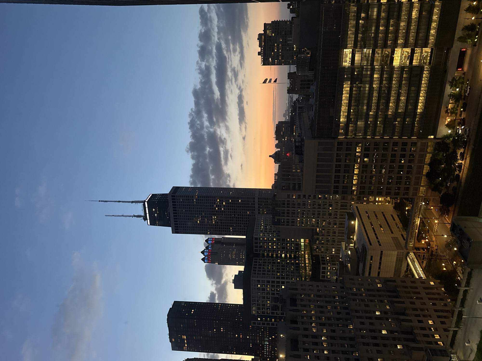 This is an aerial, vertical, full shot of the Chicago skyline at dusk. The sky transitions from a deep blue at the top to a vibrant orange and pink sunset near the horizon. Several skyscrapers dominate the scene, including the distinctive John Hancock Center, identifiable by its twin antennas. Other buildings show illuminated windows, indicating activity within. Two flags are visible on poles in the distance. The lower portion of the image shows street-level details with streetlights and parked cars. The overall impression is of a bustling urban environment at the close of day.