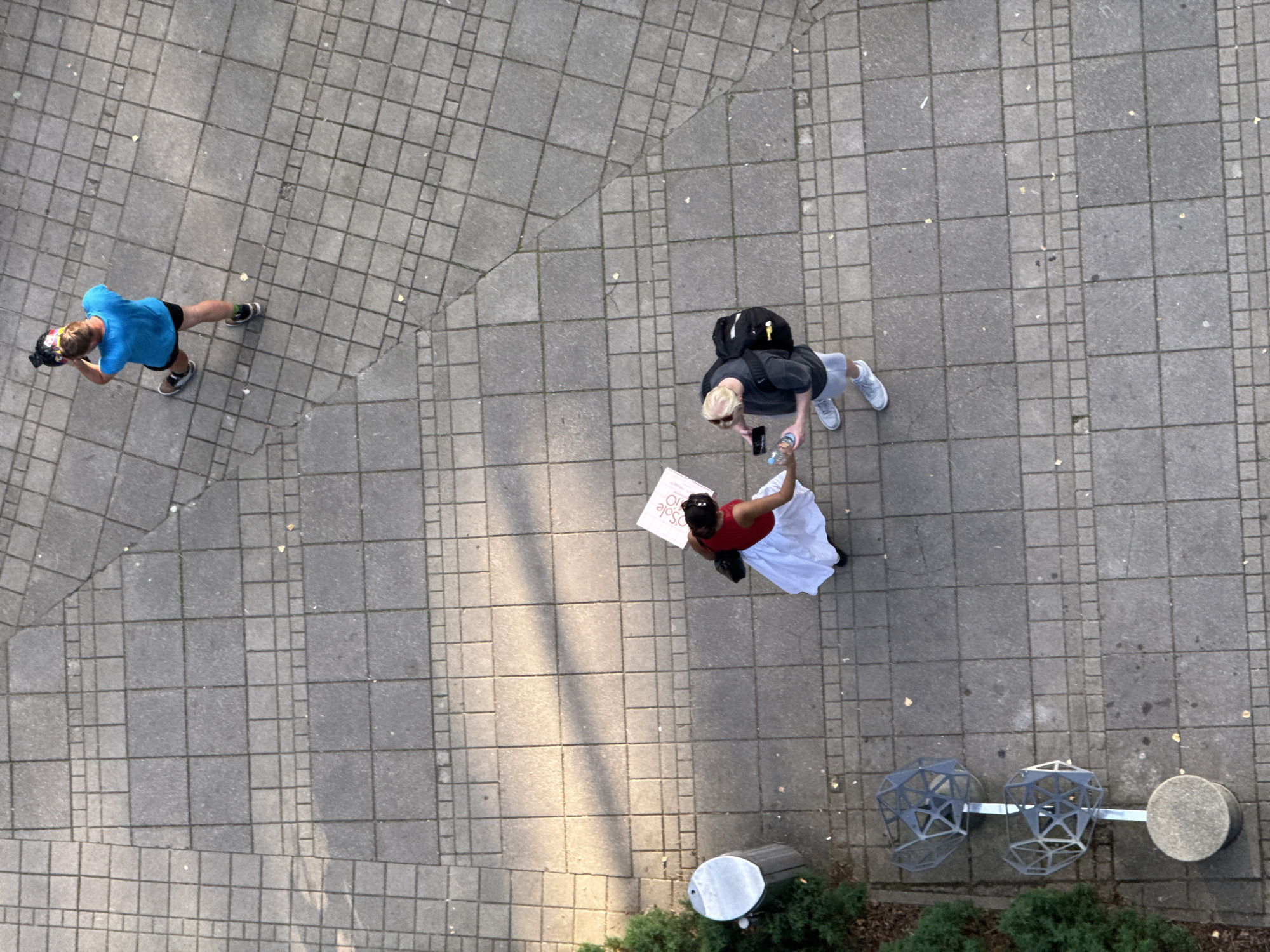 The image, taken from an elevated perspective, depicts a paved outdoor area with a pattern of interlocking square concrete tiles. In the upper right quadrant, a person wearing a bright blue t-shirt and black shorts is standing, holding a camera. In the lower half of the image, two individuals are interacting on the pavement. One, dressed in a red top and a white skirt, holds a white sign. The other, wearing a grey t-shirt, grey shorts, and a black backpack, is holding a smartphone and a bottle of water. To the left of the two interacting individuals, there is a metal trash receptacle next to some greenery. Further to the left and lower, two geometric metal sculptures are visible on a circular concrete base. The scene appears to be a public space, possibly a walkway or plaza, in Jūrmala, Latvia, characterized by its urban paving and landscaping elements.