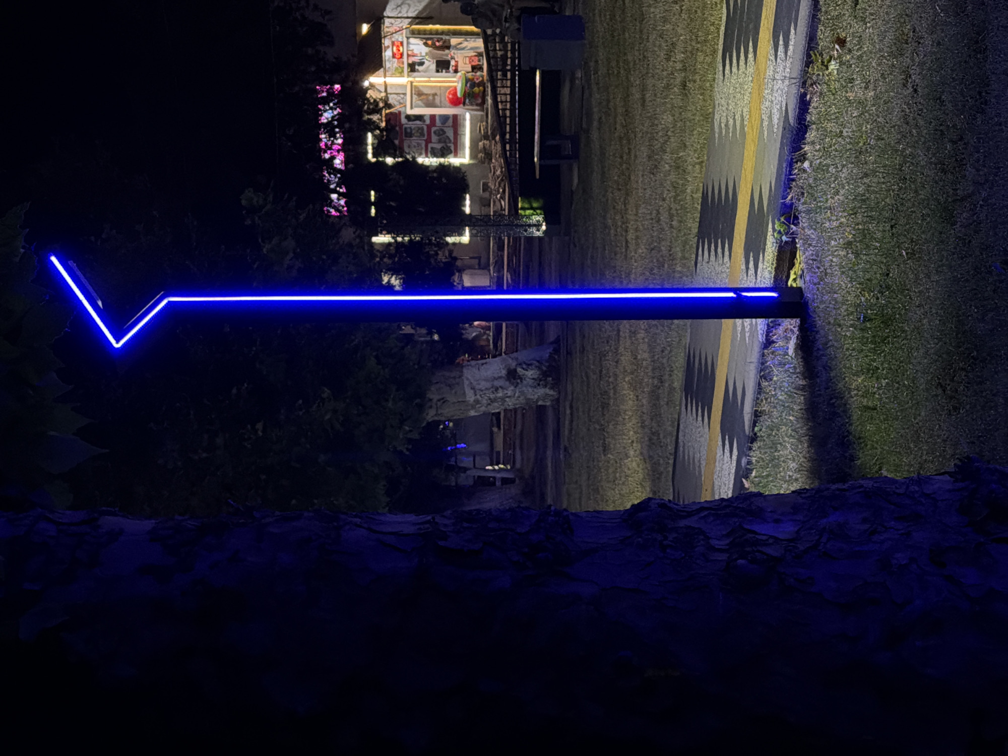 In the city of Chirchik, Uzbekistan, a street scene unfolds at night. A tall, slender lamppost with a distinctive angular, bright blue neon light fixture dominates the center-right of the frame. The lamppost is positioned on a paved pathway with a contrasting yellow and grey zig-zag pattern. To the left, the rough bark of a large tree is prominently featured, its dark silhouette partially obscuring the background. The ground is covered in dry grass, illuminated by the ambient light and the glow from the lamppost. In the background, a building with illuminated windows and colorful signage is visible, suggesting a commercial area. Benches and other street furniture are scattered around, and a few figures are present, though their details are indistinct in the low light. The overall atmosphere is one of a quiet urban evening, highlighted by the striking blue luminescence of the modern lamppost.