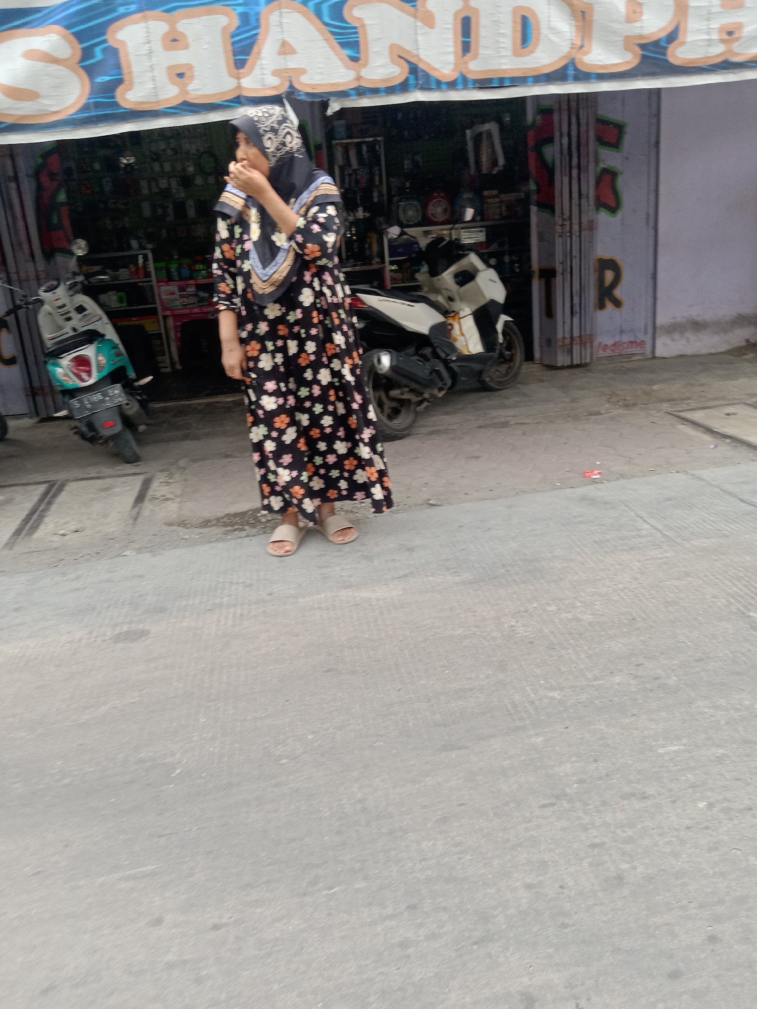 A medium shot captures a woman standing outside a store with a blue and white striped awning that reads "S...HANDPH...". The woman, wearing a black hijab and a long floral dress, stands on the right side of the frame, facing left. Her left hand is near her mouth, and her right arm is by her side. She is wearing light-colored sandals. In front of the store, two scooters are parked. A white scooter with a turquoise front is on the left, and a white and black scooter is to the right of the first scooter. The store's facade includes a metal gate with red and green graffiti and the letters "TR" visible. A small red and white object is on the ground in front of the store. The ground is paved with light gray concrete tiles. The scene appears to be on a street in Sugihwaras, Indonesia.