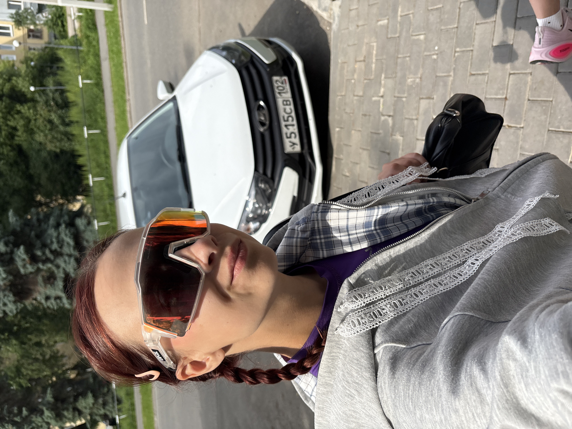 The image is a selfie taken outdoors in an urban environment, possibly in Izhevsk, Russia. A young woman with auburn hair styled in two braids and wearing oversized, mirrored sunglasses is the primary subject. She is wearing a purple t-shirt under a grey hooded jacket with white lace trim. She is holding a black purse in her right hand, which is partially visible. In the background, a white car with a Russian license plate that reads "у 515св 102" is parked on the street. The car is a Lada. The ground is paved with grey bricks. Trees and buildings are visible in the distance, indicating a city setting. A pink athletic shoe is visible in the lower right corner of the frame, suggesting another person is present but out of frame. The lighting suggests it is daytime, with sunlight casting shadows.