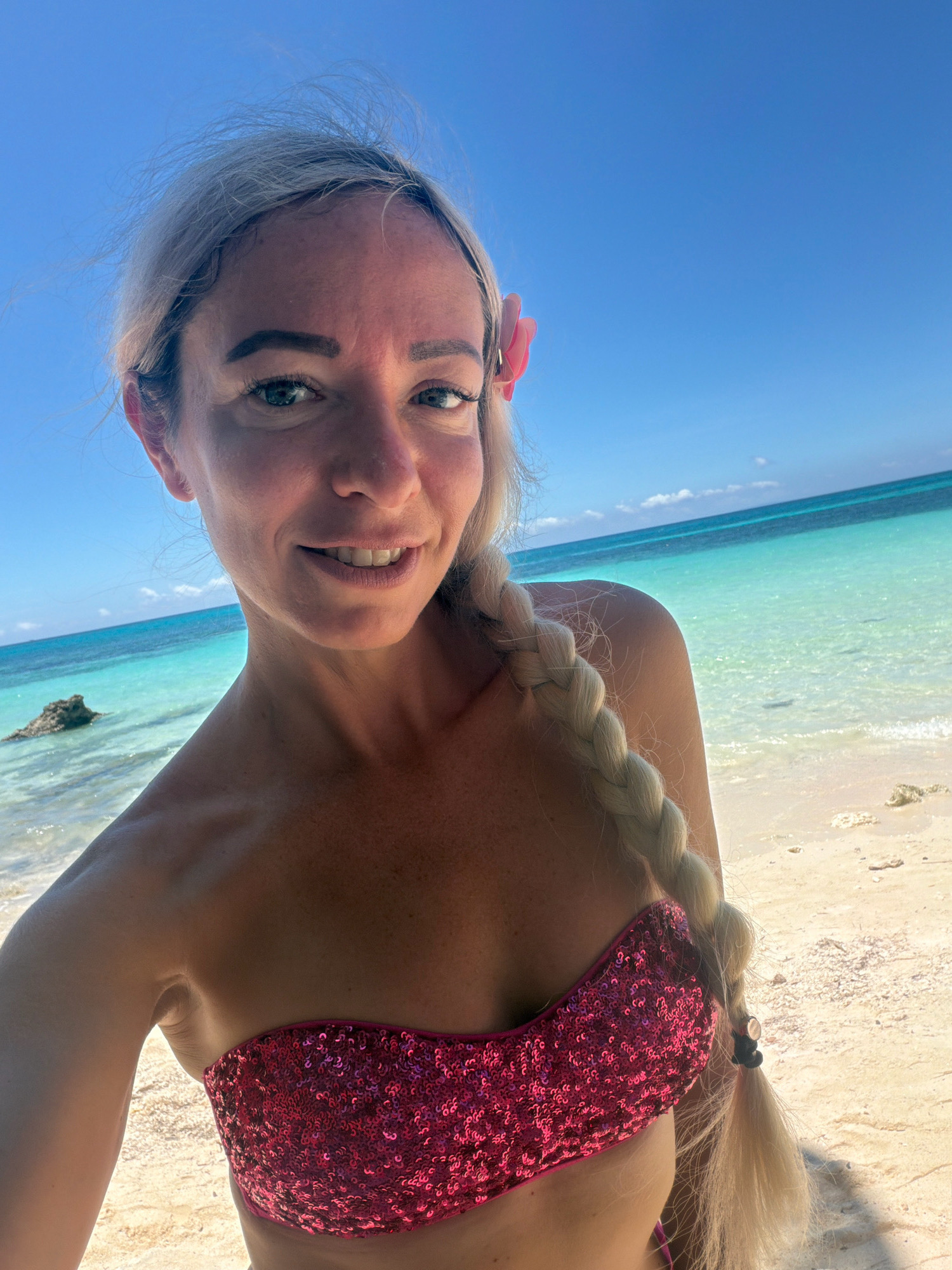 A fair-skinned woman with blonde hair braided down her back, wearing a pink sequined bikini top, is taking a selfie on a sandy beach. She has a pink flower tucked behind her left ear. The background shows a clear blue sky, turquoise water, and a rocky outcrop in the shallow sea. The location is identified as Malay, Philippines.