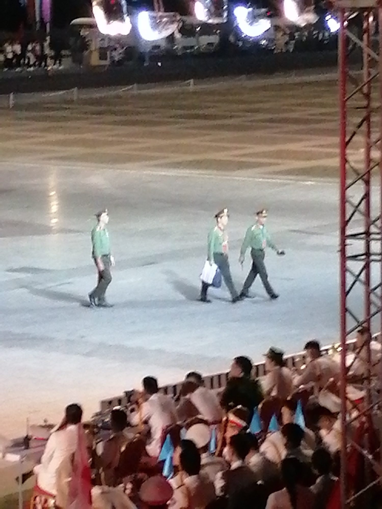 In Da Lat, Vietnam, three individuals in military-style uniforms are walking across a large, paved open area. They are wearing green shirts and dark pants, with caps on their heads. The person on the left is walking alone, while the other two are walking side-by-side. In the background, there are more people seated, some appearing to be in formal attire, and a large metal structure with red accents. The scene suggests an outdoor event or ceremony taking place in a public space.
