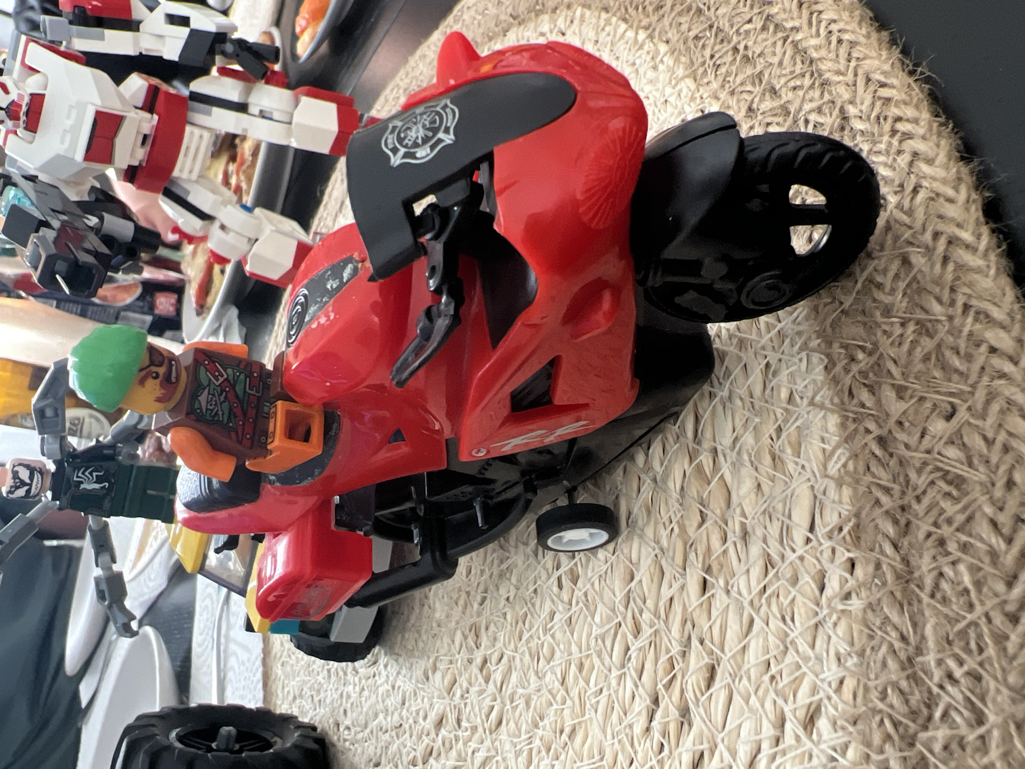The image shows a close-up view of several LEGO figures and vehicles on a woven placemat. In the foreground, a red toy motorcycle is prominently featured. A LEGO figure with a green helmet and an orange shirt is seated on the motorcycle, holding onto the handlebars. Behind the motorcycle, another LEGO figure in a dark green suit with a spider emblem is visible. To the right of these figures, a larger LEGO robot, predominantly white and red, stands. The background is blurred but suggests a domestic setting with plates of food and other objects on a table. There is no indication of any specific location in Budapest, Hungary.