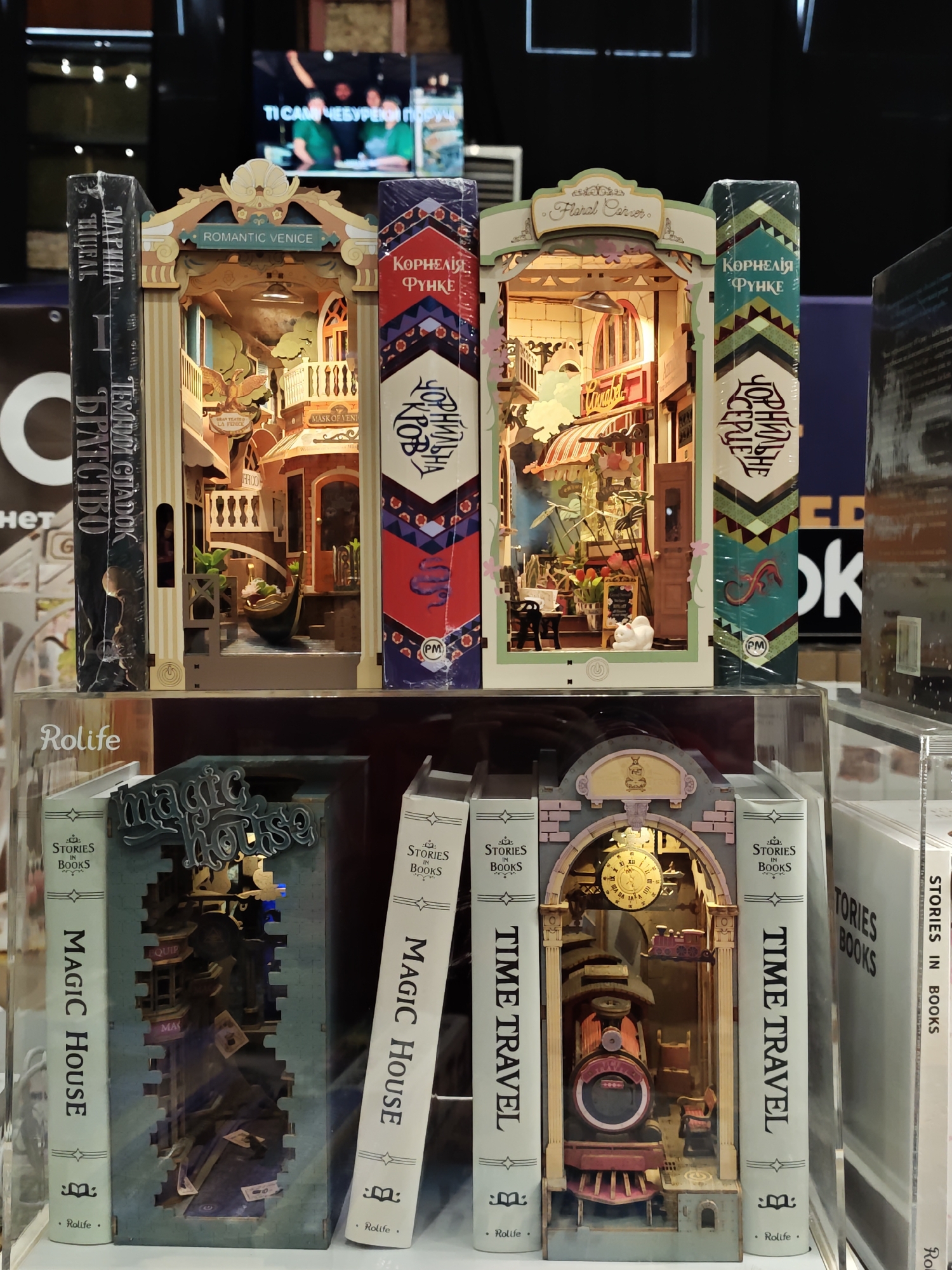 This image shows a retail display of miniature book nooks, likely a craft or hobby store, in Lviv, Ukraine. The display features several intricately designed, diorama-like structures that fit between books on a shelf.
In the upper portion of the image, three book nooks are prominently displayed. The leftmost book nook is designed to resemble a facade with the label "ROMANTIC VENICE." Inside, a scene with a gondola and architectural details is visible. To its right is another book nook, also decorated with intricate patterns, featuring the text "КОРНЕЛИЯ" and "ФУНКЕ" on its spine. This nook contains a scene with plants and small figures, possibly a floral shop. The rightmost book nook visible in this section is also a diorama, with a label that appears to read "Floral Corner." It depicts a vibrant interior with flowers and furniture.
Below these, another row of book nooks is displayed. The leftmost is labeled "MAGIC HOUSE" on its spine and features a cut-out design revealing a miniature interior scene. To its right, a book nook with the label "TIME TRAVEL" on its spine showcases a miniature train and clock face within its structure. The book nooks are branded with "Rolife."
The background suggests an indoor environment with shelves and potentially other merchandise. A large screen in the upper background displays a dimly lit video of people, though the content is indistinct. The overall scene depicts a retail setting focused on decorative book accessories.