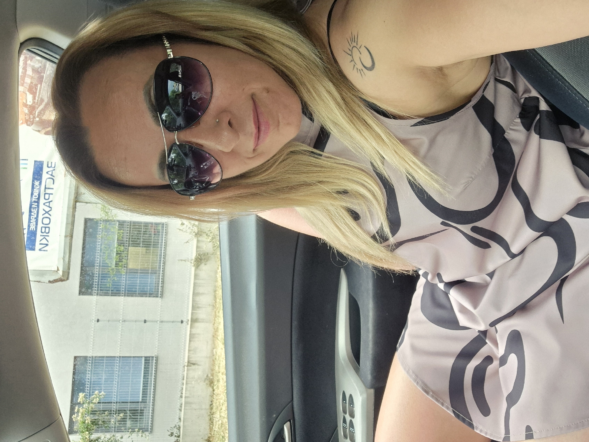 In the city of Unknown, Unknown, a woman is pictured inside a car, facing the camera. She is wearing sunglasses and a dress with a black and beige pattern. Her hair is blonde and falls over her shoulders. A tattoo of a sun and crescent moon is visible on her upper arm. The car interior, including the door panel and window controls, is visible. Outside the car, a building with windows and a fence can be seen. A billboard with text is also present.