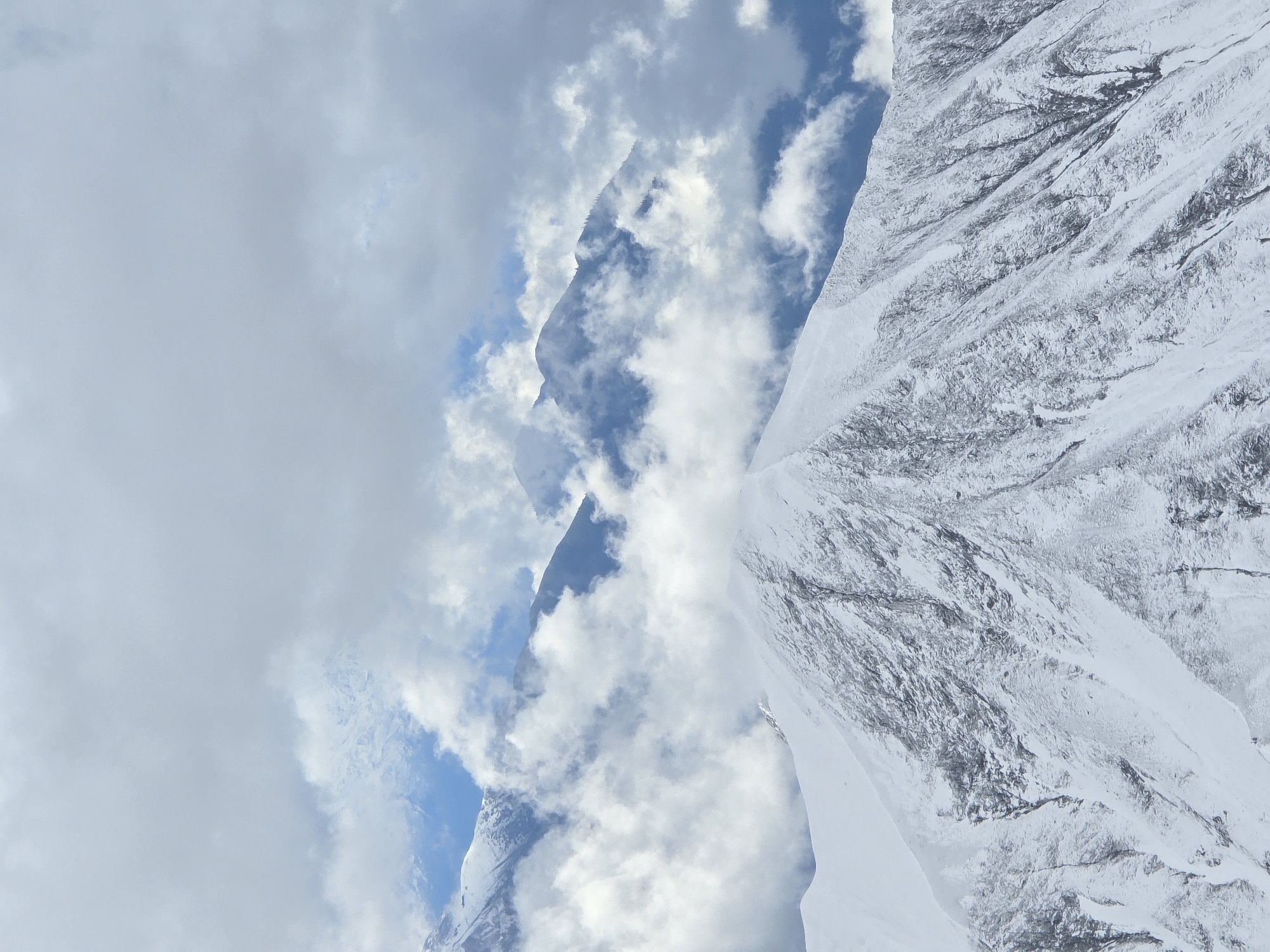 This media file depicts a dramatic, cloudy day in the mountains, likely Garmisch-Partenkirchen, Germany. The scene is dominated by towering, snow-covered mountain faces, with deep crevasses and rugged textures visible on the rocky slopes beneath the snow. Wisps of white clouds intermingle with patches of bright blue sky, creating a dynamic and slightly obscured view of the peaks.
The image is oriented vertically, suggesting it was taken from a high vantage point, perhaps looking up at the mountains or from a cable car. The heavy cloud cover indicates overcast or partly cloudy weather, with the sunlight struggling to break through. This creates a somber yet awe-inspiring atmosphere. There are no people or man-made objects visible, emphasizing the raw, untamed nature of the mountain landscape. The time of day is not clearly discernible due to the cloud cover, but the bright patches of sky hint at daylight hours. The overall impression is one of immense scale and the stark beauty of a winter mountain environment. There is no visible text in the image.