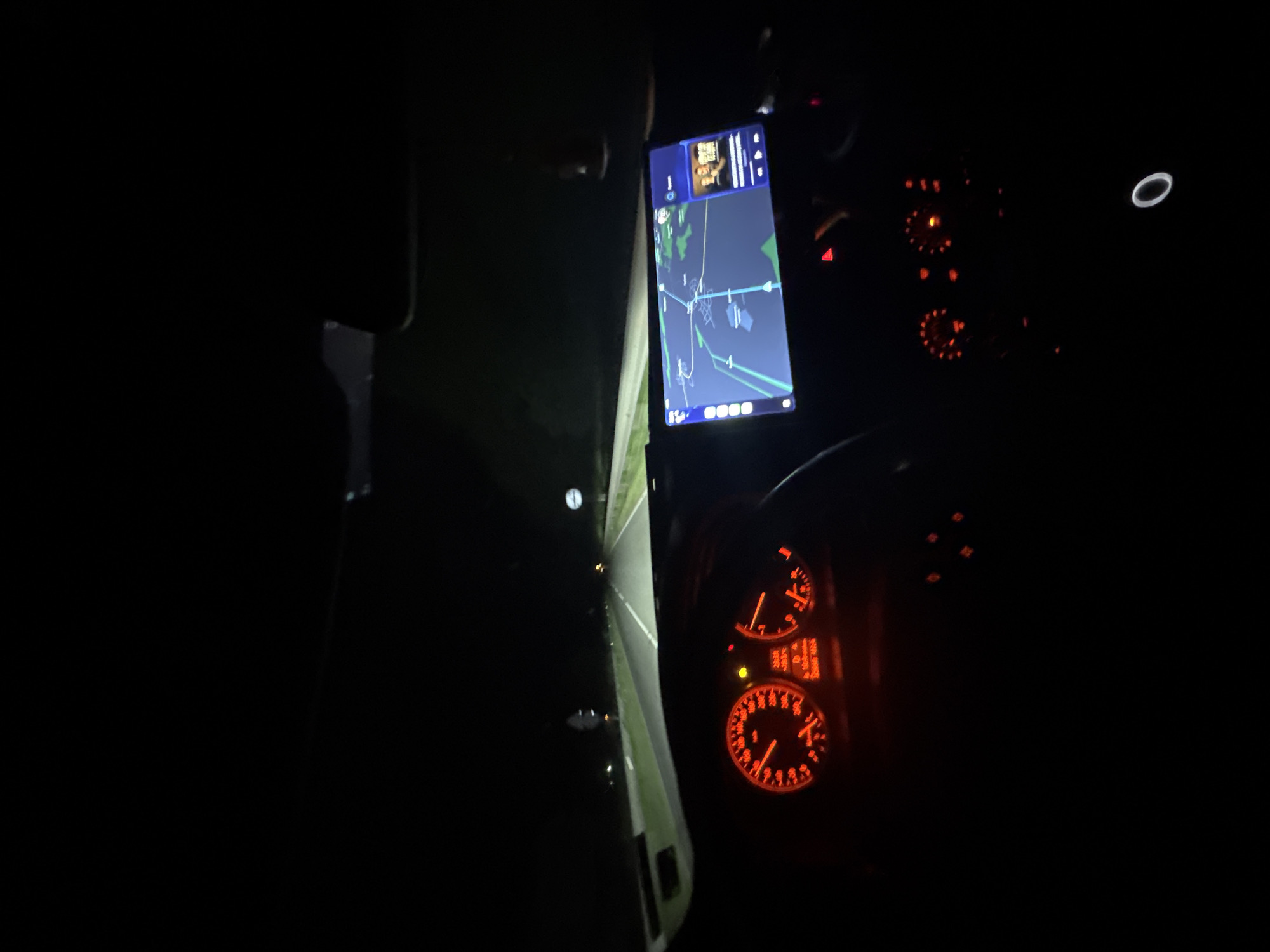 The image shows the interior of a vehicle driving at night. A navigation app is displayed on a smartphone mounted on the dashboard, showing a map with a route highlighted, potentially indicating the location in Mogila, Bulgaria. The vehicle's instrument cluster is illuminated in red, displaying gauges and indicators. The headlights illuminate the road ahead. The environment is dark, suggesting the vehicle is in an area with limited ambient lighting.