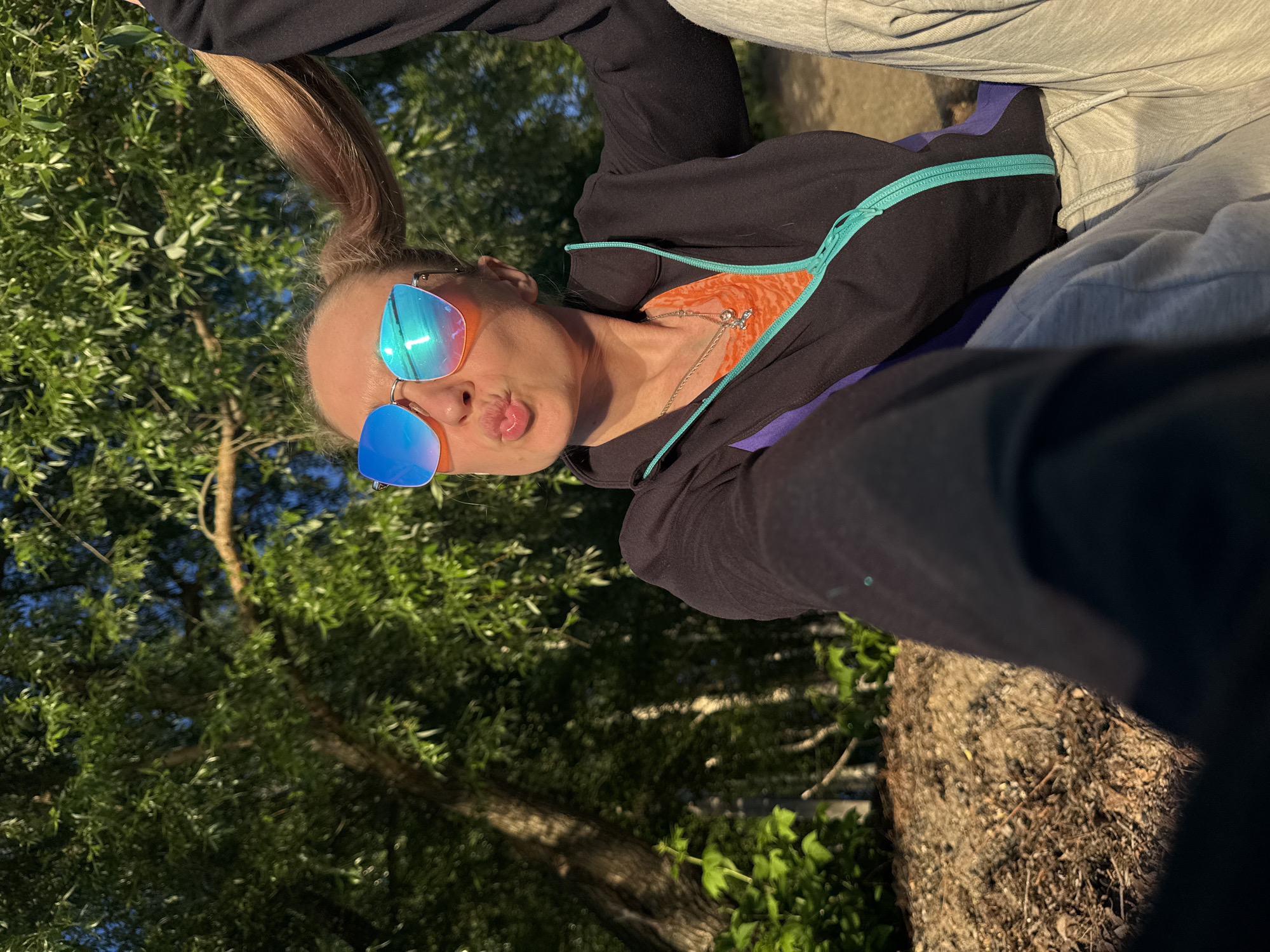 A rotated selfie captures a person in an outdoor, natural setting. The individual, likely female, has fair hair styled in a high ponytail and is making a playful "kissing" or "puckering" face towards the camera. They are wearing large, reflective sunglasses that display vibrant blue and orange hues.
The person's attire includes a black long-sleeved top or jacket with bright turquoise trim along the zipper and collar. An orange top is visible underneath the open jacket. A delicate silver necklace with a small pendant adorns their neck. The lower portion of the image shows light grey sweatpants, suggesting a casual ensemble.
The background is filled with lush green foliage from numerous trees, indicating a park, forest, or natural wooded area. Sunlight, possibly from late afternoon or early morning, filters through the leaves, creating a warm, dappled effect. The ground is a natural earthen surface, perhaps a path. The overall impression is one of leisure and cheerfulness in a natural environment. There is no visible text in the image. Given the context, this scene is set in Chaikovsky, Russia.
