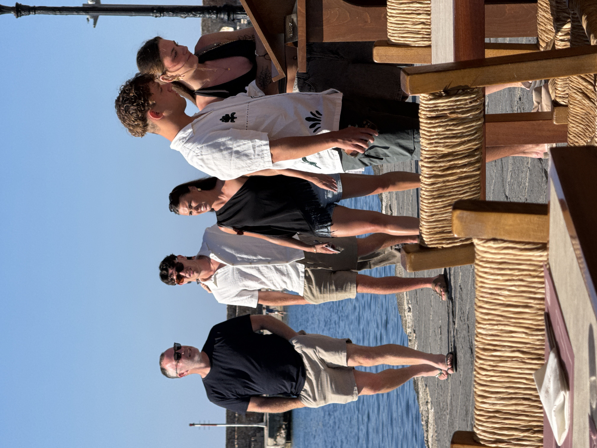 The image depicts a group of people standing on a paved walkway next to a body of blue water, likely the sea, under a clear blue sky. The walkway appears to be a pier or promenade. In the foreground, there are tables and chairs with woven seats and backs, suggesting an outdoor dining area of a restaurant or cafe.
The people are dressed in casual summer attire. On the left, a man in a black t-shirt and beige shorts is walking with his hands in his pockets. Next to him, a man in a white short-sleeved shirt and beige shorts is also seen. Further to the right, a woman in a black one-shoulder top and denim shorts is looking to her left, and a young man in a white short-sleeved shirt with a graphic print and dark shorts is looking down. To his right, a woman in a black halter top and dark shorts is looking at something on a wooden stand, possibly a menu or information board. Another person's legs are visible behind her, wearing a dark top and shorts.
The overall scene suggests a sunny day in a coastal city, possibly Chania, Greece, given the architectural style of the distant stone structures and the presence of a waterfront promenade and outdoor dining. The people appear to be casually strolling or socializing in this location.