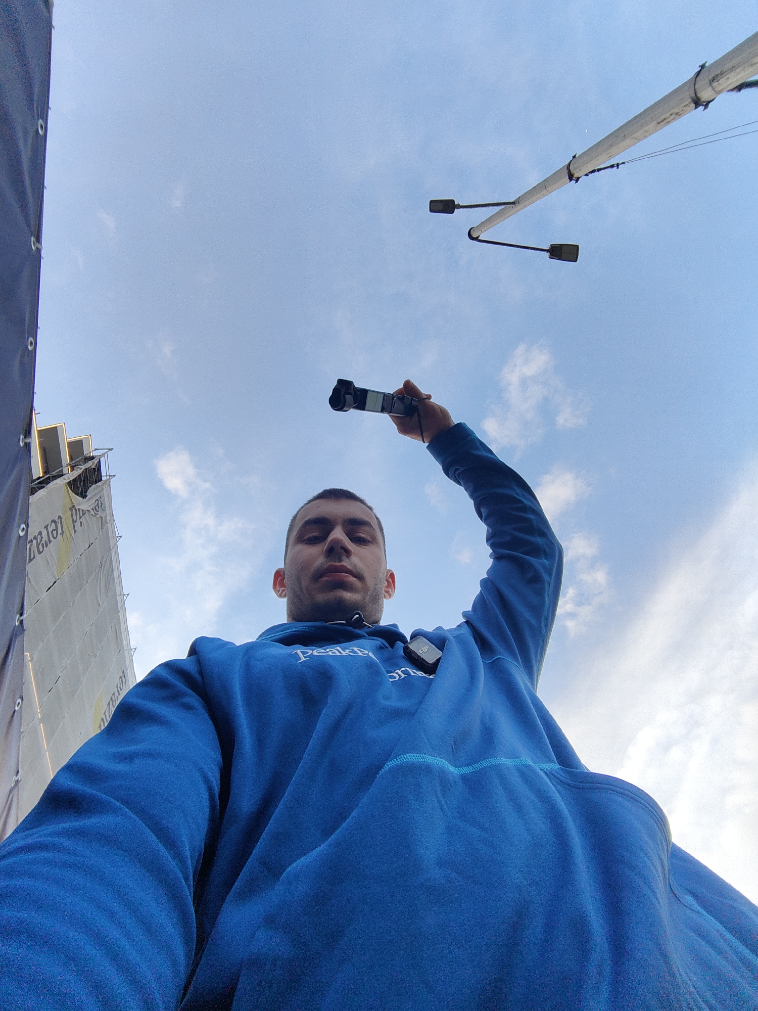 A low-angle shot depicts a man holding a camera aloft against a blue sky with scattered clouds. The man is wearing a blue hooded sweatshirt with white lettering, his arm outstretched. He appears to be recording or filming. To the left is a building facade partially covered with white material and a black tarp. Overhead streetlights are visible in the upper right corner. The location is likely Varna, Bulgaria, based on the user's prompt.