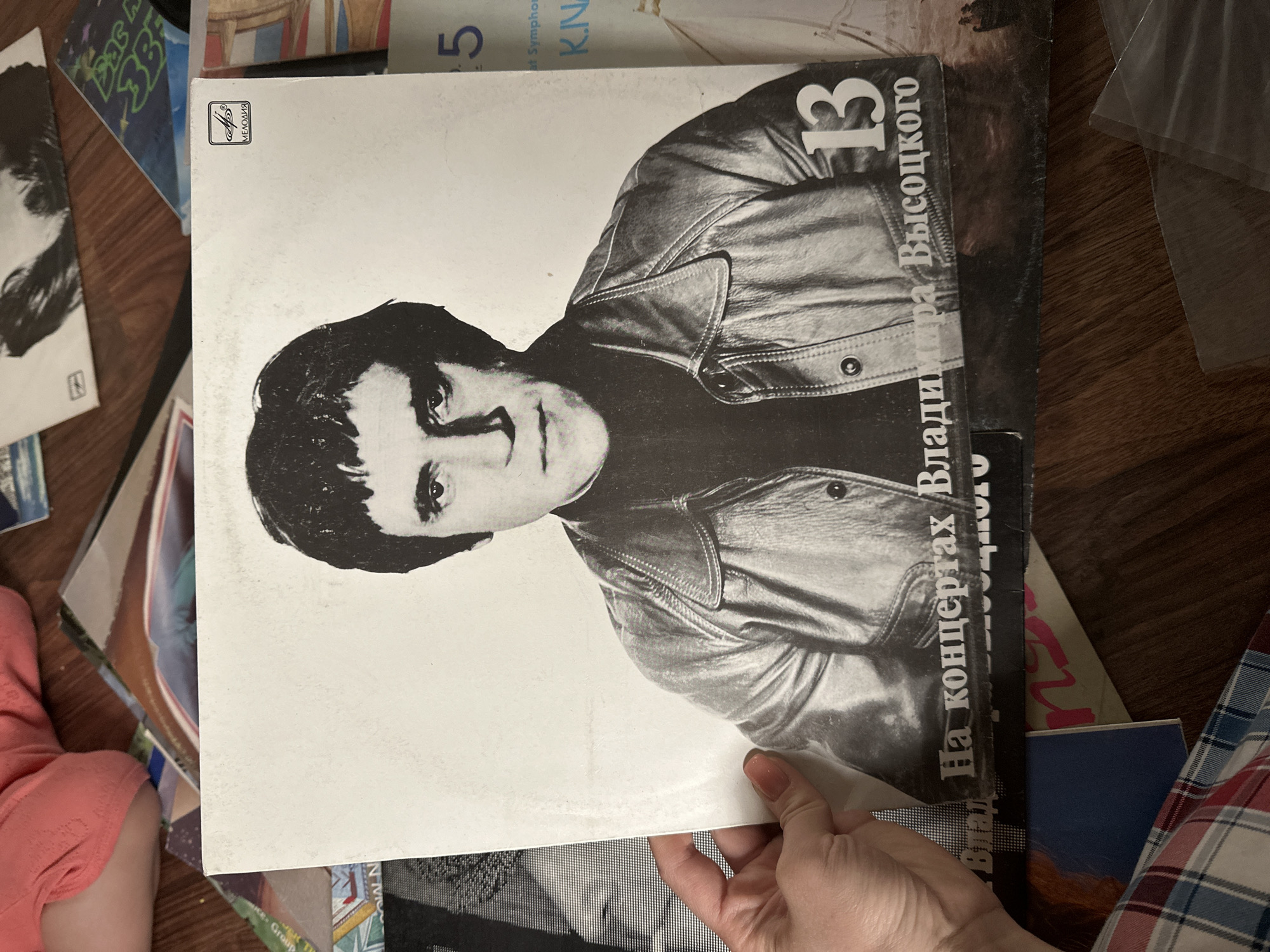 The image shows a person holding a vinyl record album of Vladimir Vysotsky, a Russian singer-songwriter and actor. The album cover is black and white and features a portrait of Vysotsky. The album is titled "13" and has Russian text written on the front. Other vinyl records and objects are scattered around the album on a wooden floor. A portion of a child's leg wearing a pink outfit is visible in the lower left corner. The location of the photo is unknown and the prompt to include Nizhny Novgorod, Russia is not applicable as there is no information in the image to indicate that location.