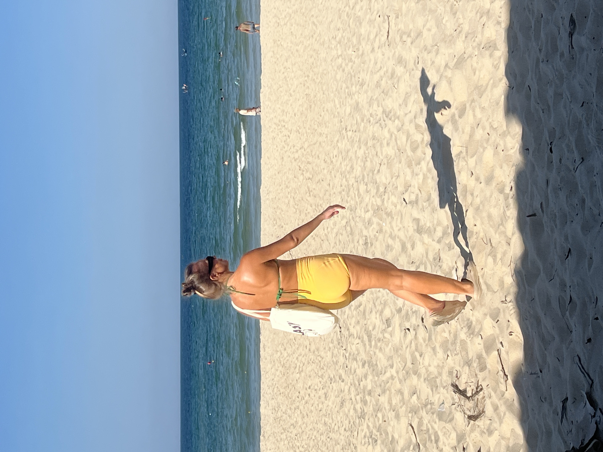 The image shows a woman in a yellow bikini standing on a sandy beach with the ocean and sky in the background. She is facing away from the camera, with her left arm extended and her right arm bent at the elbow. A white bag is slung over her shoulder. The sun is shining, casting a shadow of the woman on the sand. Several other people are visible in the distance, some in the water and some on the beach. The water is blue and there are small waves. The sky is clear and blue. The scene appears to be a typical sunny day at a beach. There is no visible text in the image.