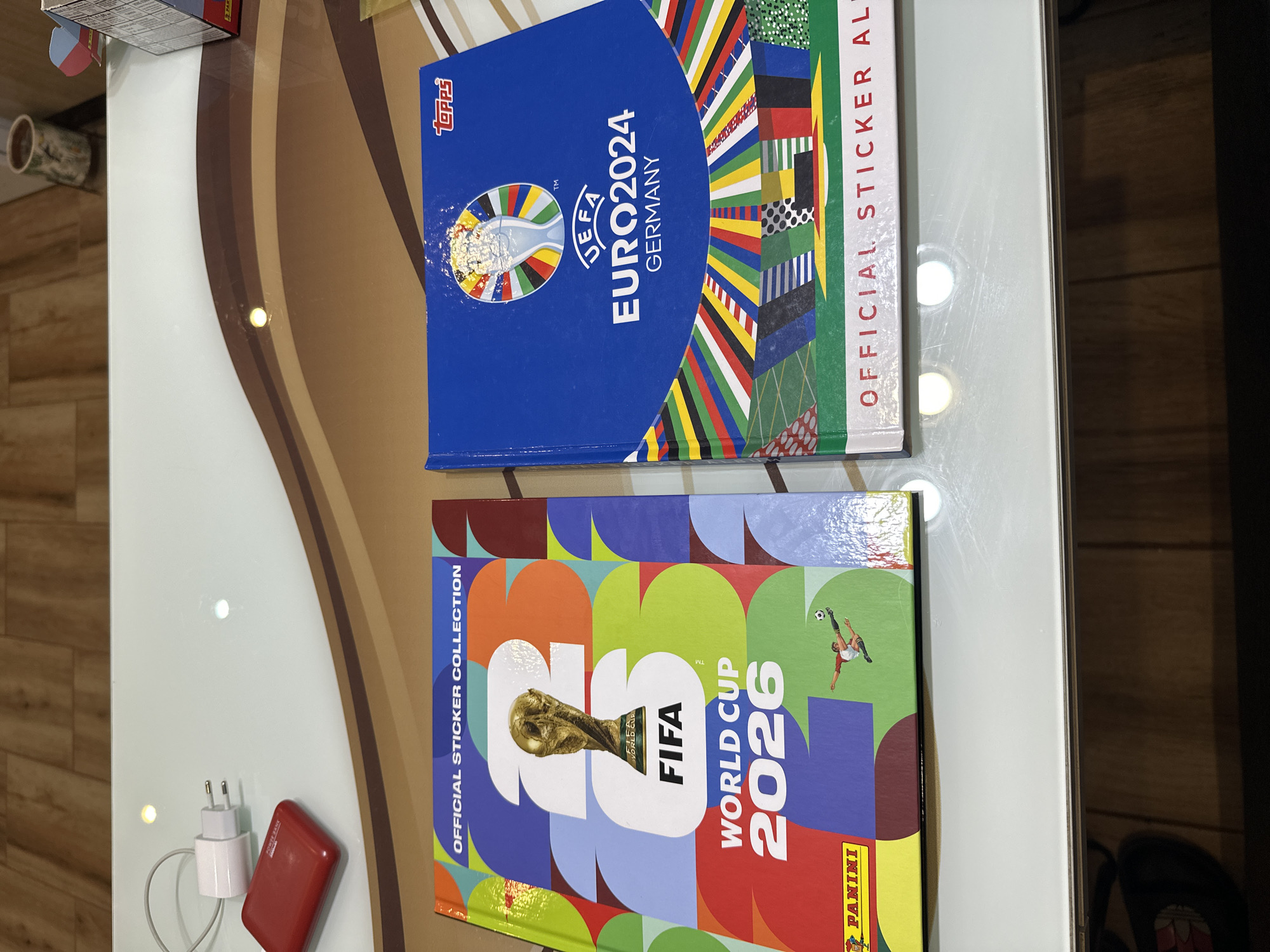 The image shows two sticker albums, one for FIFA World Cup 2026 and another for UEFA Euro 2024. The FIFA album, on the left, is predominantly colored with vibrant shapes and features a golden trophy and the FIFA World Cup 2026 logo. The UEFA album, on the right, is blue and displays the UEFA Euro 2024 Germany logo and a collage of flag patterns. Both albums are laid flat on a glass-topped table with a wooden surface visible in the background. A white electrical adapter and a red power bank are also present on the table, to the left of the albums. The scene is well-lit, with reflections from overhead lights visible on the glass surface. There is no indication of the city of Prisovo, Bulgaria, or any people present in the image.