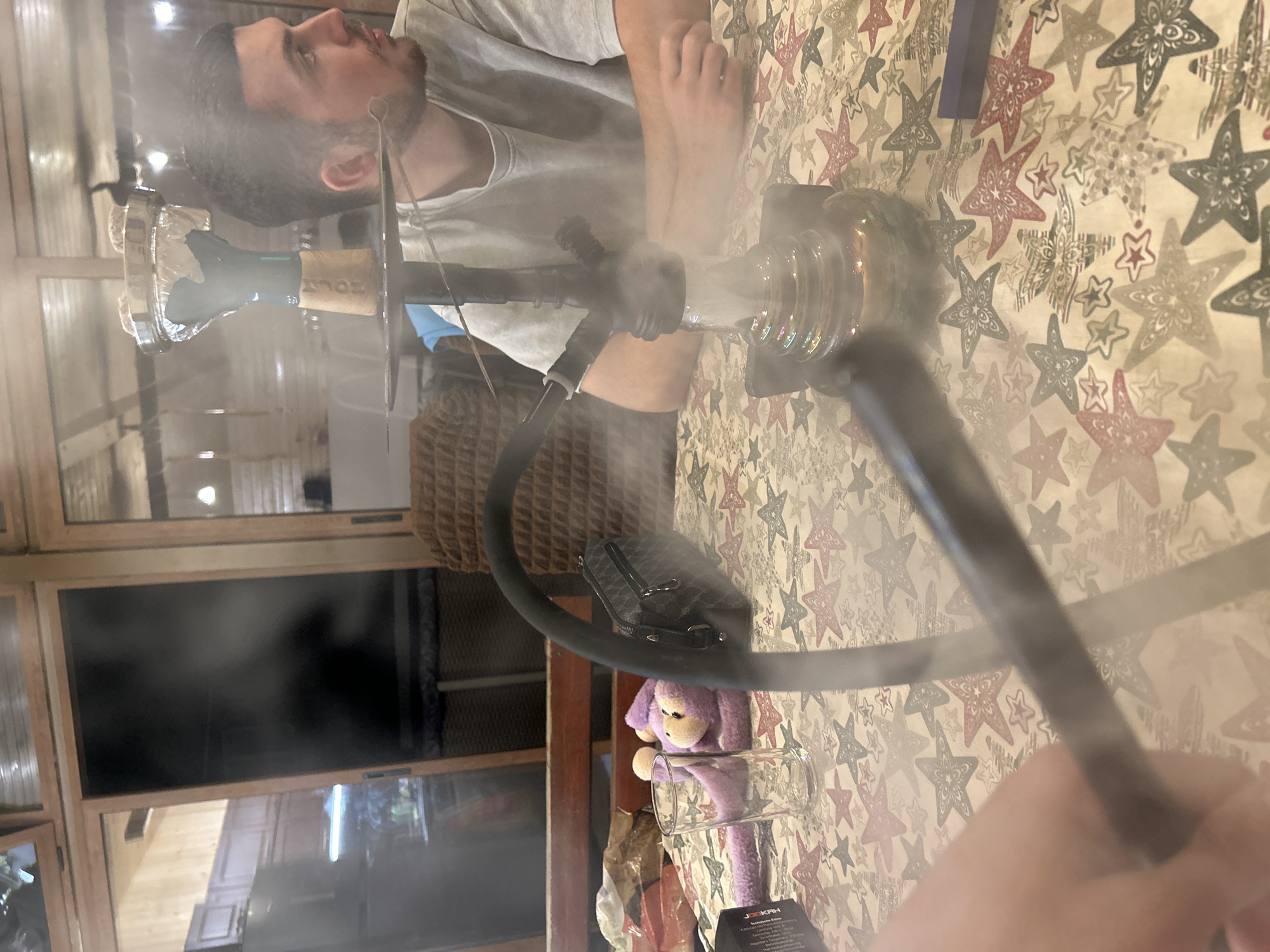 The image depicts an indoor scene, likely a residential area or a lounge, with a hookah prominently featured on a table covered with a patterned tablecloth. The table has a design of stars in red and white. A man is visible to the right, seated and looking away from the camera, with a cloud of smoke rising from the hookah. To the left, a plush purple monkey toy and a clear glass are placed on the table, along with a black handbag. The background shows wooden paneling and furniture, suggesting a rustic or cozy interior. The lighting is diffused, and the presence of smoke adds an atmospheric element to the scene. There is no specific indication of the city of Каменар, Bulgaria, within the visual information provided.