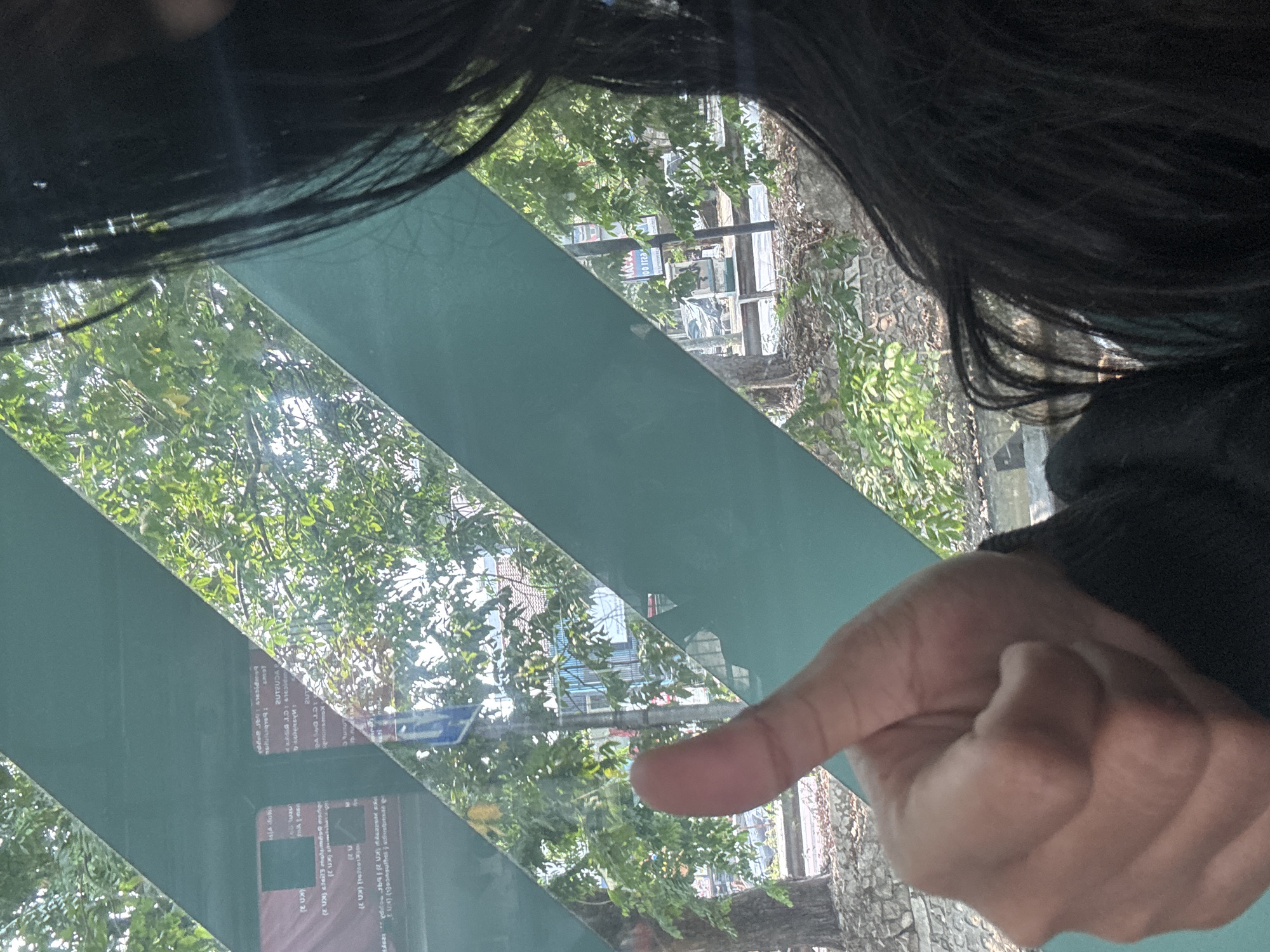 A person's left hand, with the thumb extended upwards in a "thumbs up" gesture, is visible in the foreground, obscuring a portion of the background. The hand is positioned in front of a green, slanted reflective surface, possibly a window or partition. Beyond this surface, lush green trees and foliage are visible, suggesting an outdoor environment with natural light. In the distance, there are hints of urban elements such as buildings and possibly a street. The overall impression is that the photograph was taken from inside a vehicle or a building with a view of a street scene. The location in North Jakarta, Indonesia is not discernible from the visual information provided in the image.