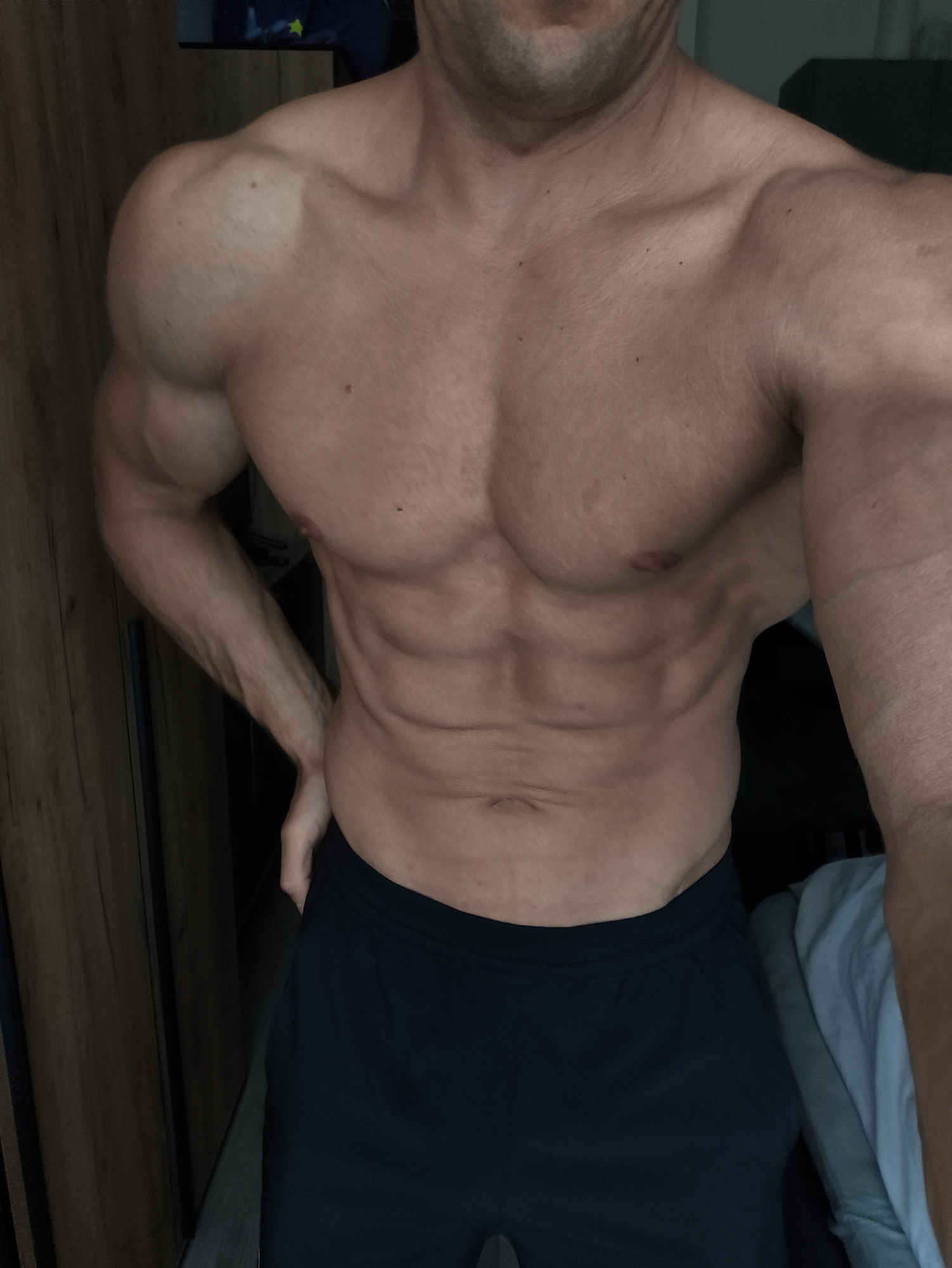 The image features a shirtless male individual, cropped from just below the chin to the upper thighs. The subject exhibits a highly muscular and lean physique, with clearly defined pectorals, a visible six-pack abdomen, prominent obliques, and well-developed shoulders and arms. Veins are visible on the forearms, indicating low body fat. Several small, dark moles are scattered across the torso. The individual's left hand is placed on their hip, suggesting a posed stance. Dark blue or black athletic shorts cover the lower portion of the body.
The scene is an indoor setting with subdued, uneven lighting. To the left, a section of a warm brown wooden wardrobe or cabinet with vertical grain is visible. To the right, a light blue or grey fabric object, possibly bedding, is partially visible, with a darker, indistinct background behind it. The upper background behind the subject's head is dark and lacking specific features. There are no elements within the image that provide information about its location in Moscow, Russia.