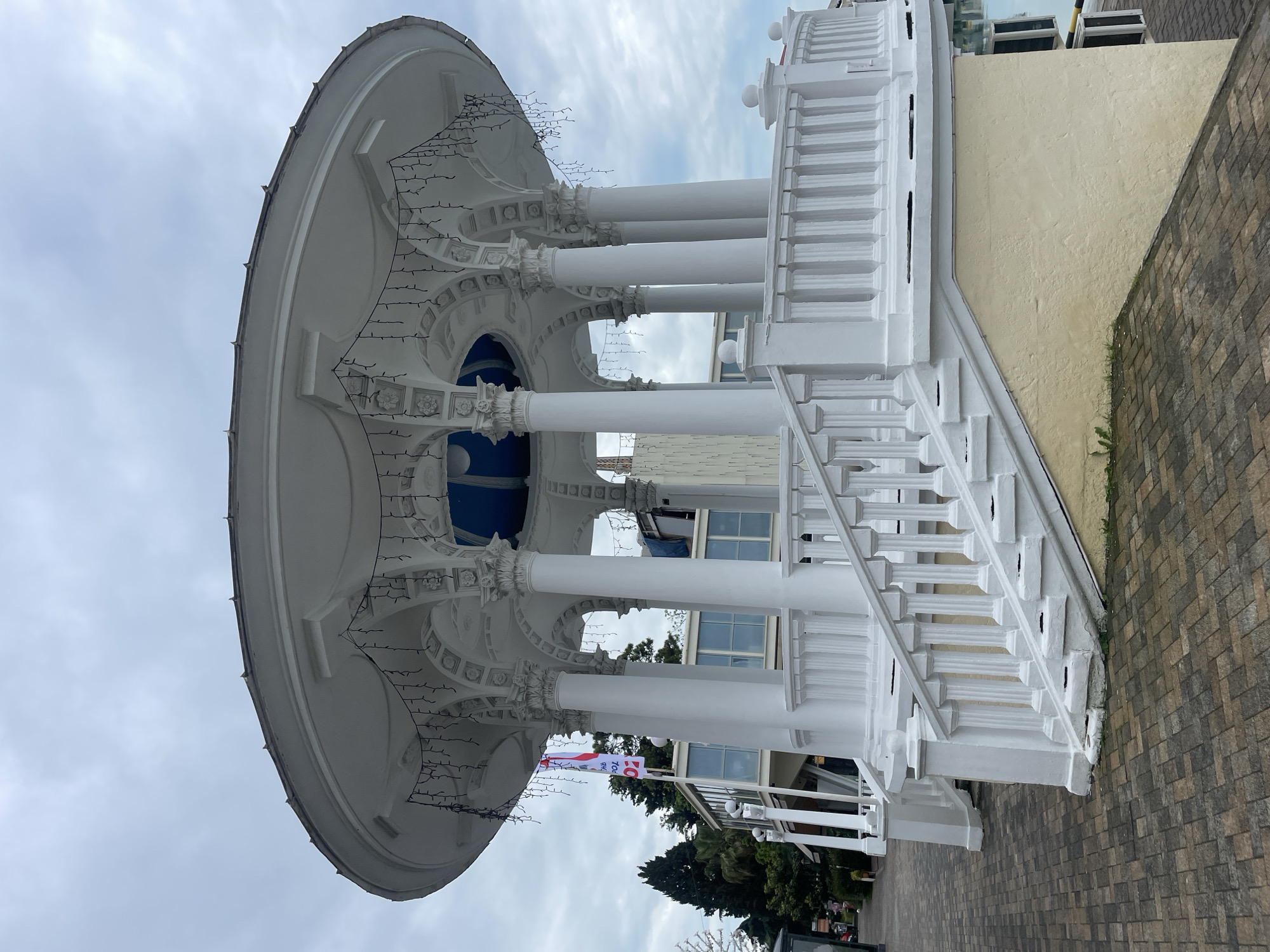 The image shows the rotunda pavilion on the Sochi embankment, Russia, a white structure with cylindrical columns, a domed roof, and decorative lighting. The structure has a staircase with a balustrade. A flag is visible near the base of the pavilion. The background includes trees, a building with windows, and a cloudy sky. The surrounding area is paved with brick tiles.