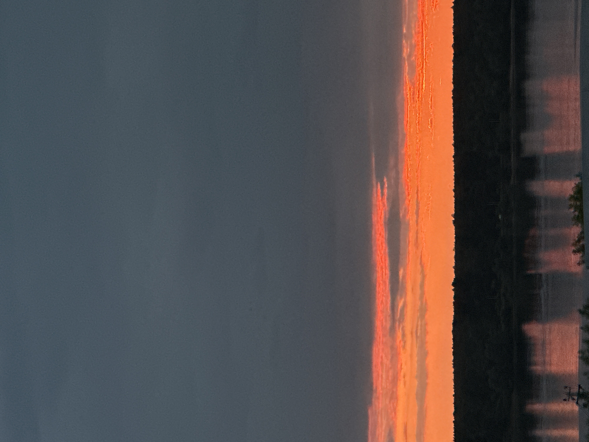 This is a vertical, full-frame photograph of a sunset over a body of water, likely the Kama River, near Chaikovsky, Russia. The sky is dominated by dark, moody clouds in the upper half of the image, transitioning to vibrant orange and pink hues near the horizon. A thin band of clouds reflects the intense sunset colors. Below the sky, a dark silhouette of a forest lines the far bank of the river. The river itself is calm, reflecting the colors of the sunset and the dark treeline, creating shimmering streaks of light on its surface. In the foreground, a dark, indistinct line suggests the edge of the land or a structure. The overall atmosphere is serene and dramatic, capturing the fleeting beauty of twilight.