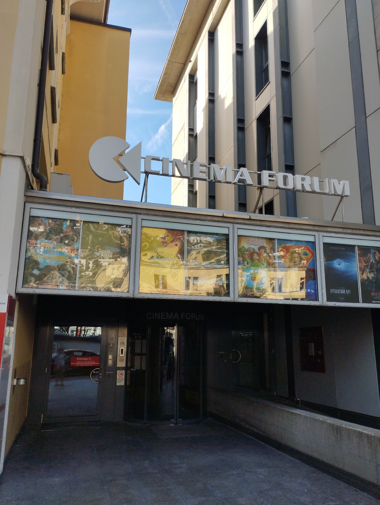 An outdoor, eye-level shot captures the entrance of a building prominently displaying "CINEMA FORUM." The cinema entrance is recessed, featuring a dark, paved ramp leading to automatic glass doors. Above the entrance, a horizontal array of lightboxes showcases various movie posters, including titles partially visible like "HEADS WILL ROLL," "SCARY MOVIE," "Minions," "SUPERGIRL," "DISCLOSURE DAY," and "SOLO NETEREMT" (as per OCR).
The cinema's main signage, "CINEMA FORUM," is in large, white, three-dimensional letters above the lightboxes, accompanied by a distinct white logo resembling a Pac-Man character. The building housing the cinema is modern, with light grey panels and dark grey vertical beams. To the left, a different building with a yellow facade and cream-colored sections is visible.
The sky is clear blue with faint wispy clouds, indicating daytime, and shadows are cast by the buildings, suggesting sunny weather. Reflections of the surrounding buildings and a red "Entrata &" (Italian for "Entrance") sign, along with smaller text "Fabbrocini - Cassa del Popolo" and "UFT Cinema Forum T.3", are visible on the glass doors. Faint reflections of people are also discernible on the glass and poster displays. No direct human activity is clearly depicted, though the scene implies a functional cinema. A concrete barrier runs along the right side of the ramp leading to the entrance.