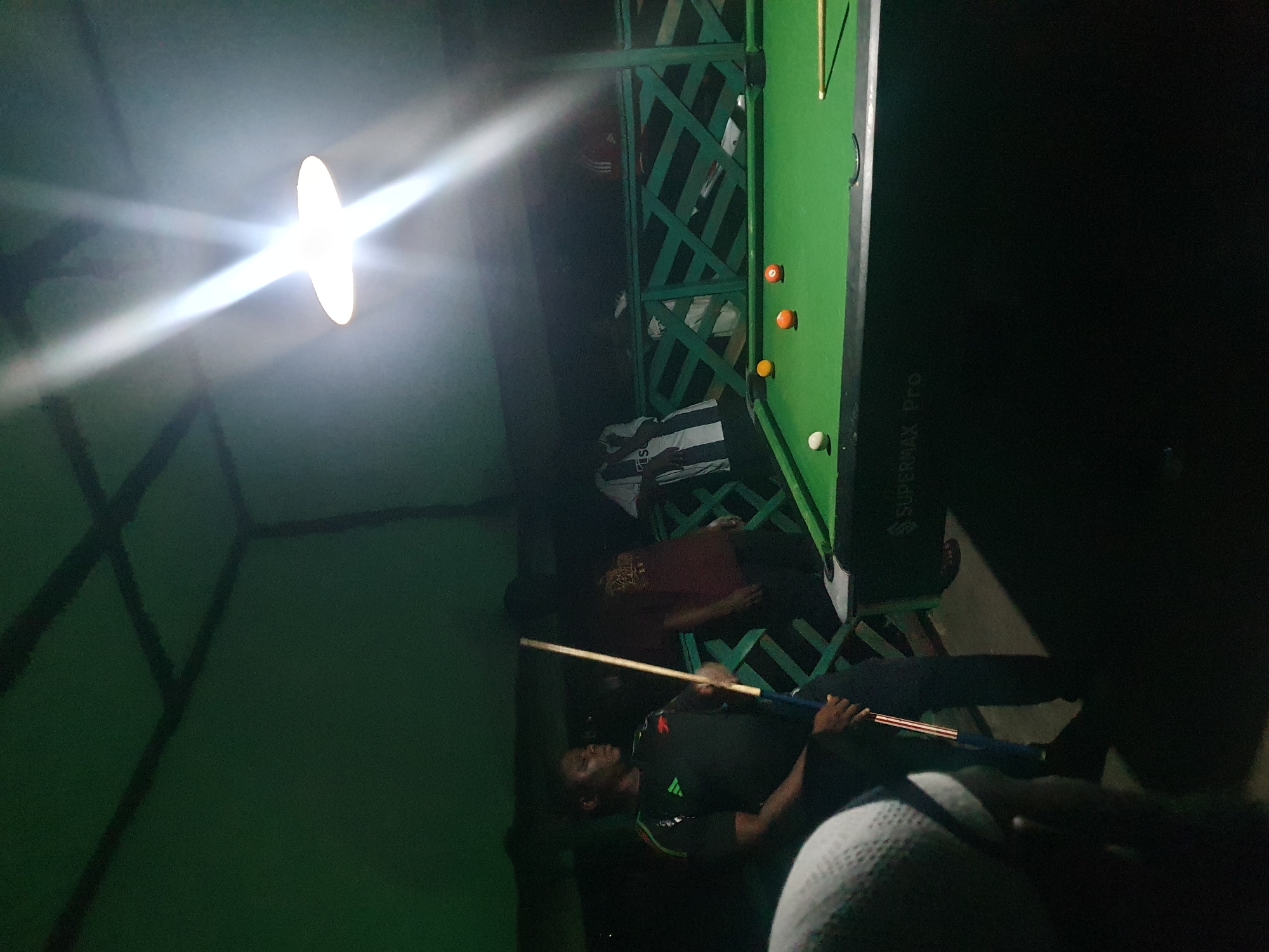 The image depicts a group of people playing pool at night, possibly in an outdoor covered area. A pool table with green felt is visible, with several billiard balls on it, including an orange one, a yellow one, and a white one. Two men are standing near the table. One man, wearing a dark t-shirt with colorful stripes on the sleeves, is holding a pool cue and appears to be in the process of playing. Another man in a maroon t-shirt stands behind him. The environment is dimly lit, with a bright overhead light casting strong beams. The background shows a dark green, slatted fence or railing. The scene suggests a casual recreational setting. The presence of a "SUPERMAX Pro" inscription on the pool table indicates it is a professional or commercial grade table. While the specific location within Lokoja, Nigeria, cannot be determined from the image alone, the scene is consistent with a local sports bar or outdoor entertainment spot in the city.