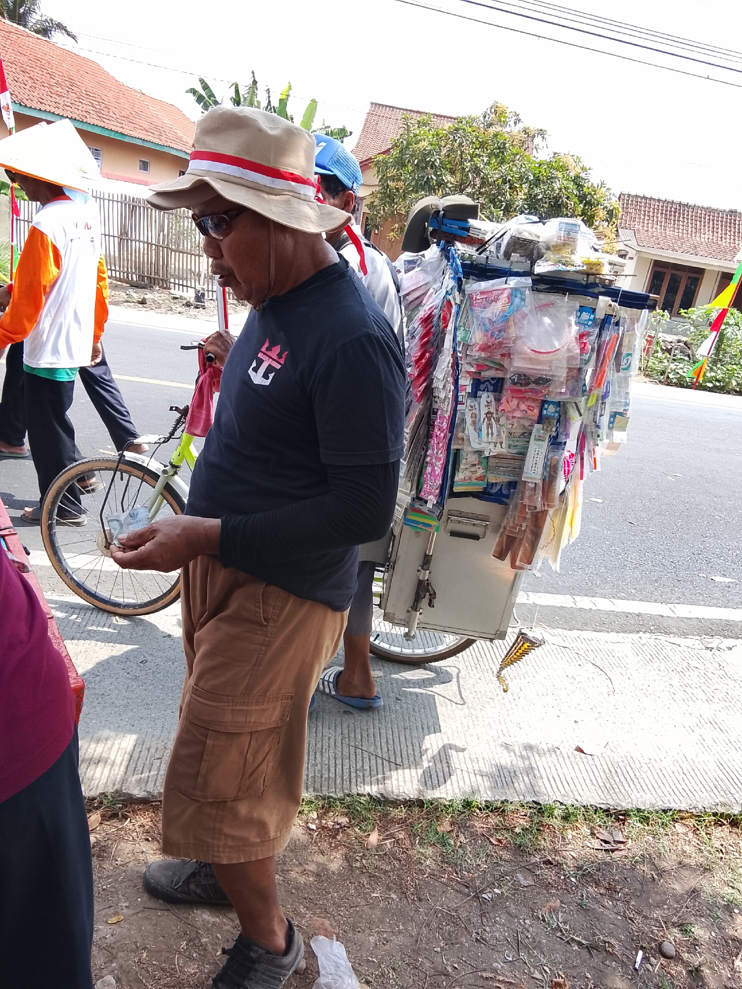 In Sampang, Indonesia, on a bright day, a man wearing a tan hat with red and white bands, sunglasses, and a dark t-shirt stands by a bicycle loaded with various packaged items, possibly for sale. He is holding some Indonesian Rupiah banknotes in his hand. Behind him, other individuals are visible, one in an orange and white shirt with a conical hat, and another with a blue hat. The bicycle's cart is laden with an assortment of merchandise, displayed in clear plastic packaging and hanging from a rack. The scene is set on a paved roadside with a concrete sidewalk adjacent to a dirt and grass area. Buildings with orange-tiled roofs and greenery are in the background. The sun casts shadows, indicating it is daytime.