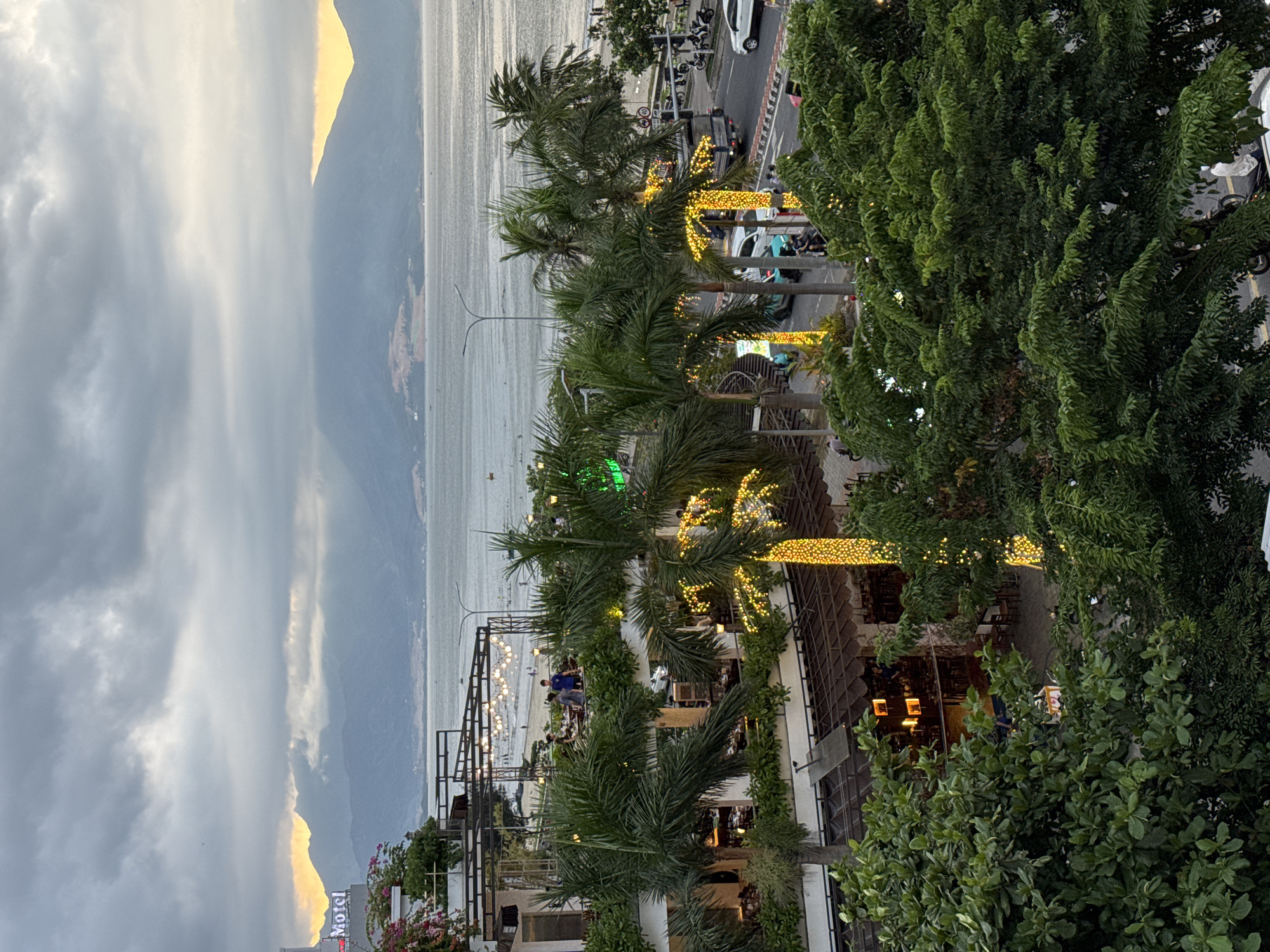 This image captures a view of the beachfront in Đà Nẵng, Vietnam, likely near the Sơn Trà district, characterized by its numerous resorts and restaurants lining Võ Nguyên Giáp Street. The scene depicts an overcast sky with muted sunlight breaking through the clouds, illuminating the distant mountains. In the foreground, a multi-story establishment with an open-air restaurant and balconies is visible, adorned with palm trees wrapped in decorative yellow lights. Several people are seen dining or walking on the balconies. Beyond the building, a beach with scattered individuals and beach umbrellas stretches towards the sea. A road runs parallel to the beach, with a few vehicles, including cars and motorbikes, present. The overall atmosphere suggests a late afternoon or early evening setting, with the artificial lights on the palm trees beginning to illuminate the scene.