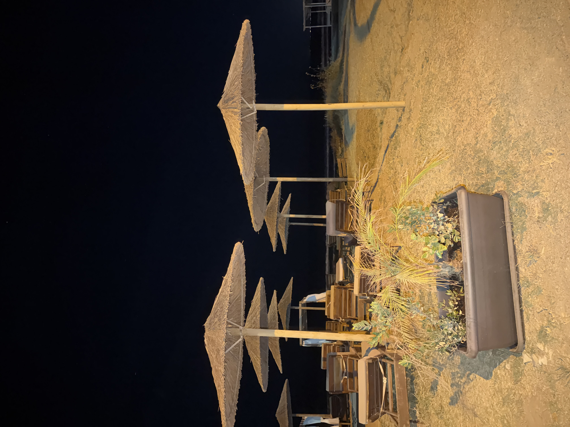 The image depicts a beach scene at night in Georgioupoli, Greece. In the foreground, a large rectangular planter contains various green plants, including what appears to be a small palm frond. Behind the planter, several rows of empty wooden lounge chairs are neatly arranged on a sandy surface. Above the chairs, multiple thatched umbrellas are propped up by wooden poles. The sky above is dark, indicating nighttime, with a few faint stars visible. The overall atmosphere is quiet and serene, with artificial lighting casting a warm glow on the chairs and umbrellas.