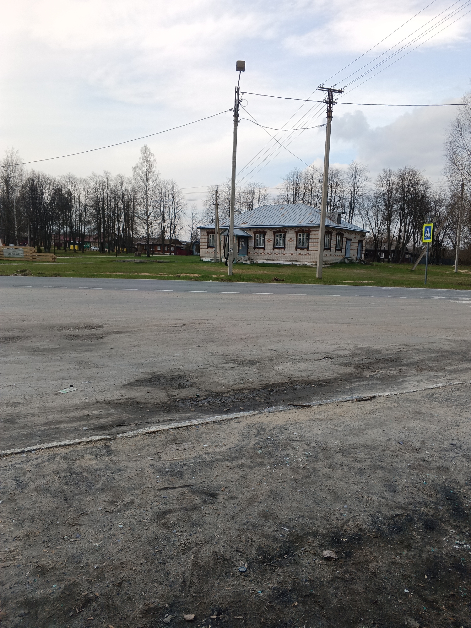 The image depicts an unpaved roadside in Красный Маяк, Russia. The road is cracked and patchy, bordering a grassy area. Beyond the grass is a single-story brick building with a gray roof, situated among bare trees. Power lines and poles run alongside the building. A yellow pedestrian crossing sign is visible on the right. The sky is overcast.