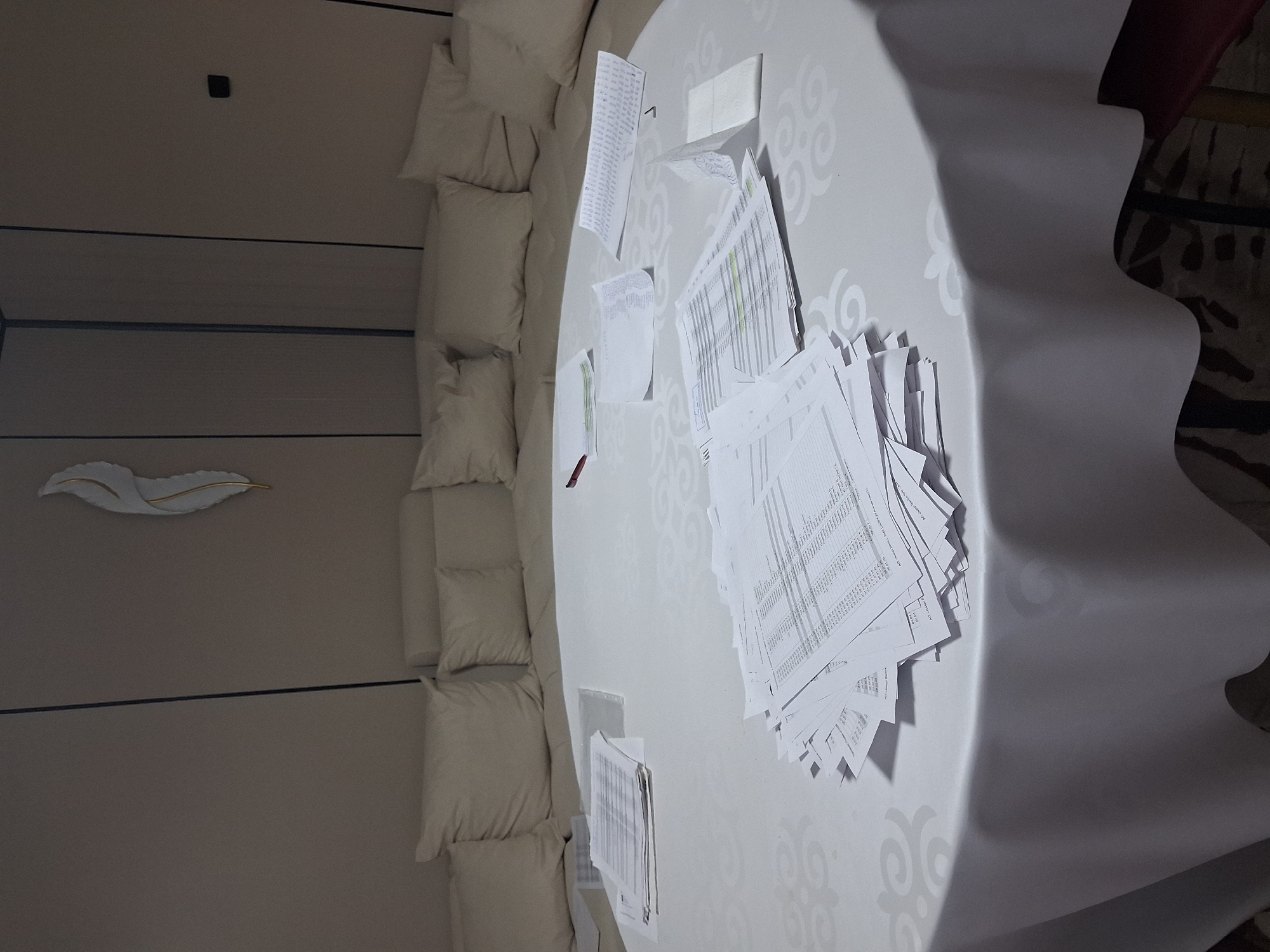The image depicts a round table covered with a white tablecloth, laden with scattered stacks of papers, some neatly organized and others appearing disheveled. The papers seem to be documents with text and numerical data, suggesting a workspace or a meeting setting. The table is placed in a room with a beige upholstered sectional sofa in the background. A decorative feather-like object is mounted on the wall above the sofa. The lighting is subdued, casting a soft glow on the scene. The location is identified as Guldala, Kazakhstan. There are no individuals present in the image, and no discernible actions or interactions are taking place. The overall impression is one of a temporarily abandoned work area.