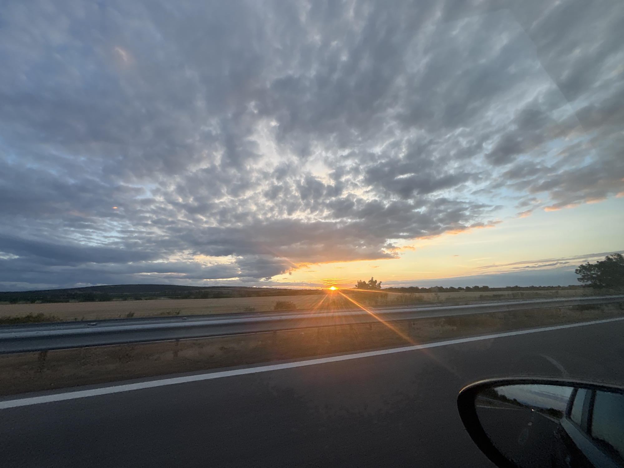 The image depicts a road scene during sunset, with the sun setting on the horizon. The location is identified as being in the vicinity of Shtarkovo, Bulgaria. A highway with a white line marking the lane is visible in the foreground, with a metallic guardrail and a concrete barrier to the left. Beyond the road, there is a field of dry grass and rolling hills covered with vegetation under a cloudy sky. The sunset is casting a warm glow, with visible sun rays emanating from the setting sun, which is partially obscured by clouds. The reflection of the sky and the surrounding landscape is visible in the side mirror of a car, which is positioned in the lower right corner of the frame. There are no people or animals clearly visible in the scene. The overall atmosphere is one of a tranquil rural landscape at the end of the day.
