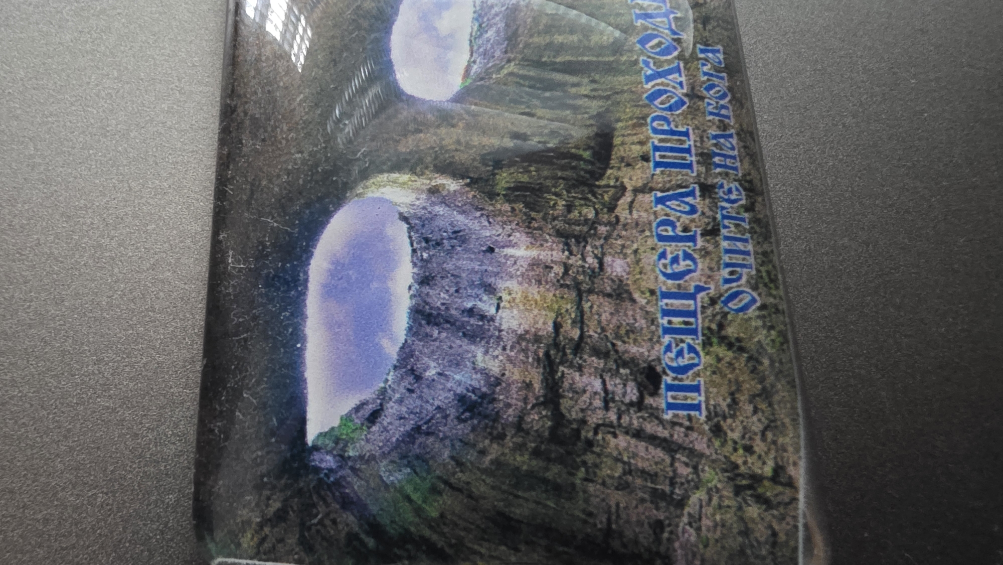 The image shows a vertical photograph of a postcard or similar item, featuring a photograph of the God's Eyes cave system in Blagoevgrad, Bulgaria. The cave system is characterized by two large openings in the cave's ceiling, allowing daylight to enter. The openings are oval, and the light is visible through them. The rock formations surrounding the openings are rugged and textured, with a mix of brown, grey, and hints of green. To the right of the image are Cyrillic letters, likely the name of the location or some descriptive text. The postcard has a glossy finish, indicated by the glare in the top left corner. The background is a solid, dark gray, possibly the surface where the postcard is placed.