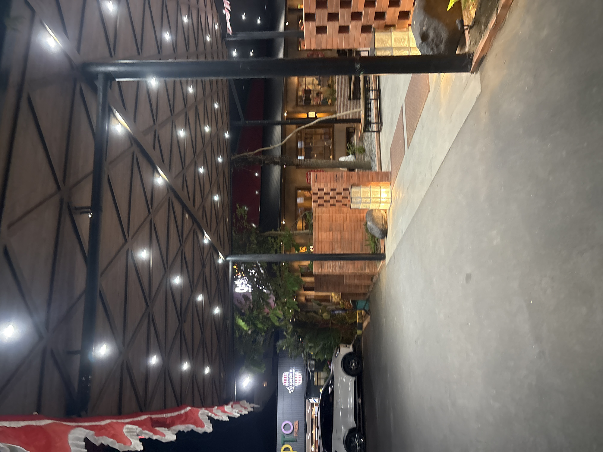 The image shows an exterior view of a modern commercial establishment at night, located in Cirebon, Indonesia. The building features a triangular-patterned roof with embedded lights, supported by dark structural beams and pillars. In the foreground, a paved walkway leads towards the establishment. To the left, a white SUV is parked. The establishment itself is characterized by brick walls, some of which are constructed in a lattice pattern, and illuminated glass block structures. Trees and foliage are visible within the property. Signage for "PTO PARK" and "COFFEE BARREL" is present on the building's facade. The overall atmosphere is well-lit, suggesting a commercial area open for business.