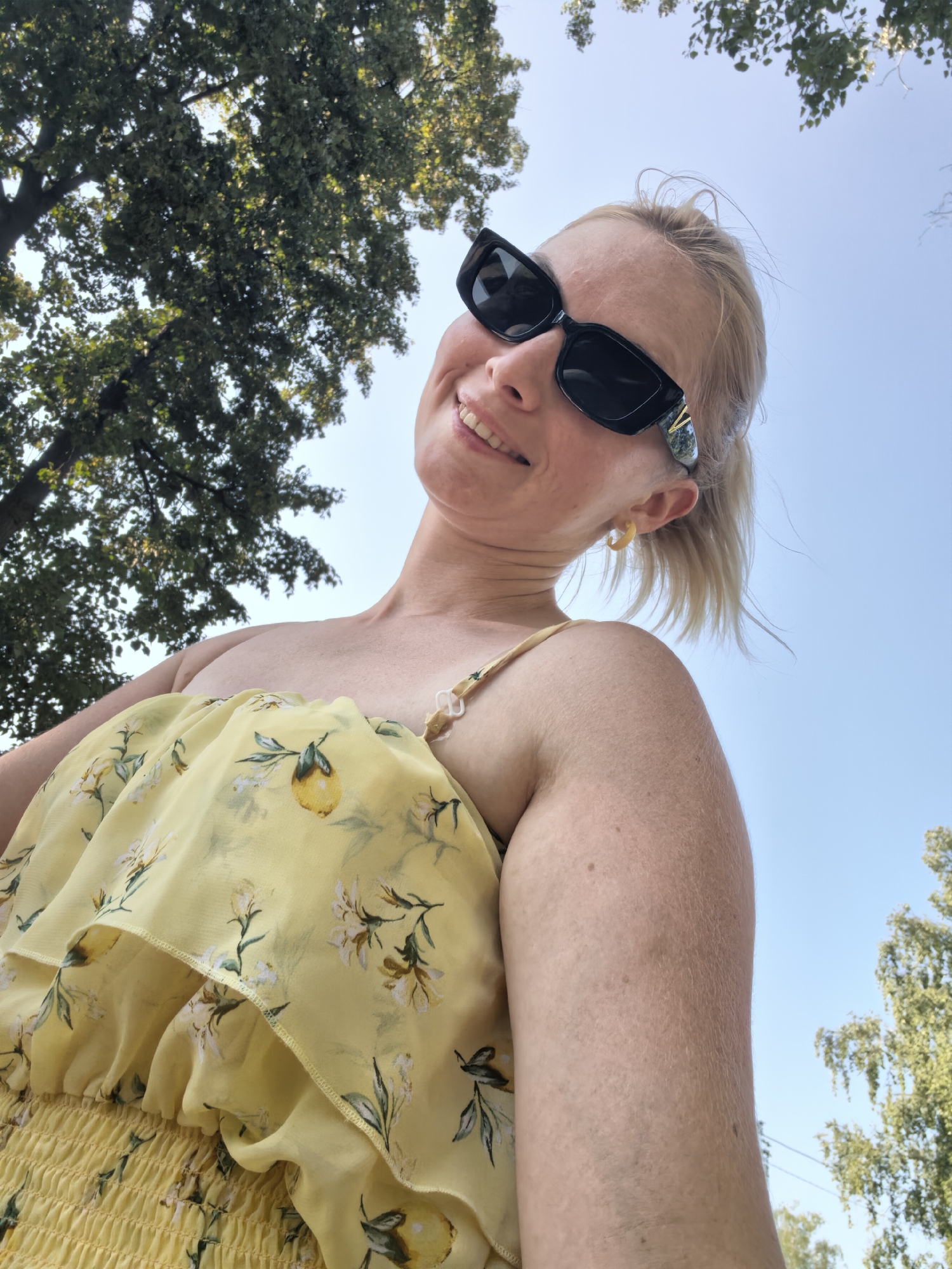 In an outdoor, low-angle shot, a fair-skinned woman with blonde hair pulled back in a ponytail is looking up at the camera with a smile. She is wearing black cat-eye sunglasses, a yellow dress with a lemon and floral print, and a light yellow hoop earring in her visible ear. The dress has a ruffled overlay and smocked fabric at the bodice. The background consists of a clear blue sky with the tops of green trees visible in the upper left and right corners. The setting appears to be a park or natural area, possibly in Sterlitamak, Russia, on a bright, sunny day.