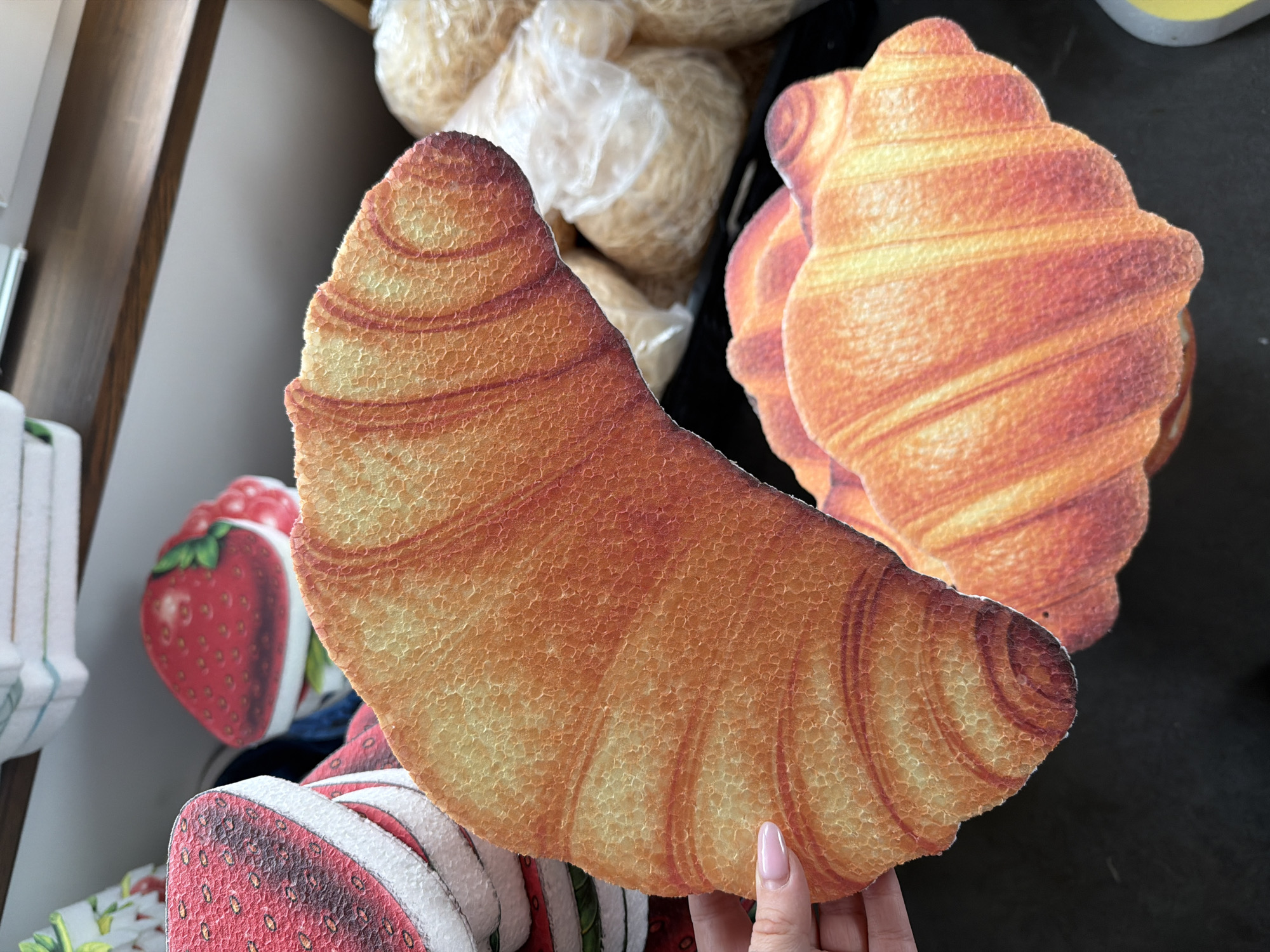 In Ternopil, Ukraine, a close-up shot reveals a dark surface with several artificial food items. In the foreground, a hand with a light pink fingernail holds a large, crescent-shaped cutout resembling a croissant, its textured surface detailed with shades of orange and yellow. Partially visible beneath it is another similar cutout, also depicting a croissant. To the left, a stack of red and white circular cutouts, designed to look like strawberries, are stacked, with one clearly showing a strawberry pattern. In the background, several translucent plastic bags are filled with what appears to be shredded or thin pasta. The overall scene is an indoor setting, likely a display or storage area.
