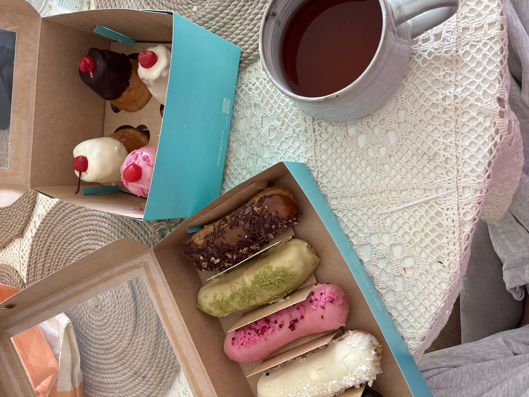 The image depicts a flat-lay arrangement of desserts and a drink. A light-colored mug filled with a dark liquid, possibly tea, sits on a white, crocheted doily. Two cardboard boxes are positioned near the mug. One box, a turquoise color, contains four round pastries, each with a different frosting and topped with a cherry. The other box, a brown cardboard color with a clear window, contains four eclairs, each with a different glaze. The location is most likely Krasnoyarsk, Russia, based on the inscription on the turquoise box.