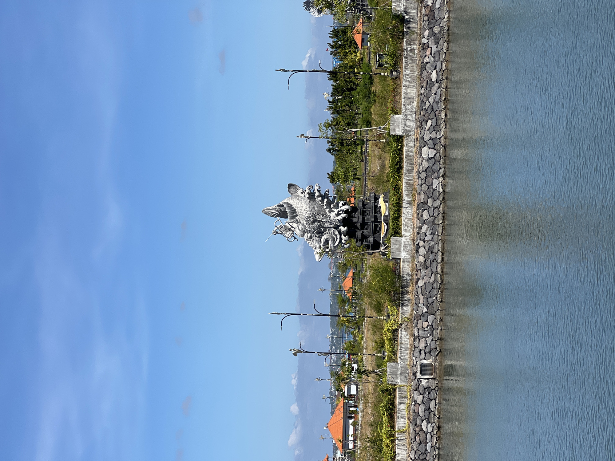 The image captures a view of the **Garuda Wisnu Kencana Cultural Park** in South Denpasar, Indonesia. The prominent subject is the partially visible, colossal statue of **Garuda**, a mythical bird-like creature, which is a key element of the larger Garuda Wisnu Kencana statue complex. The statue, rendered in a metallic, possibly stone or concrete, material, features intricate details of feathers and a dynamic posture. It is positioned on a raised platform with a concrete retaining wall and a base constructed from dark, rough-hewn rocks.
In the foreground, a body of calm, blue water reflects the sky and the surrounding environment. The water's surface shows slight ripples. Behind the statue and the rocky embankment, the park landscape unfolds. This includes patches of dry grass, scattered green shrubs and trees, and several ornamental lampposts with decorative elements.
Further in the background, there are structures with distinctive orange-tiled roofs, characteristic of Indonesian architecture. These buildings appear to be part of the park's facilities or surrounding areas. Beyond these, a hazy horizon shows more trees and buildings, suggesting an urban or semi-urban setting. The sky is a bright, clear blue with scattered, wispy white clouds. The overall scene is bathed in bright daylight, indicating a sunny day. There are no people visible in the image. The atmosphere is serene and monumental, highlighting the scale and artistic merit of the statue.