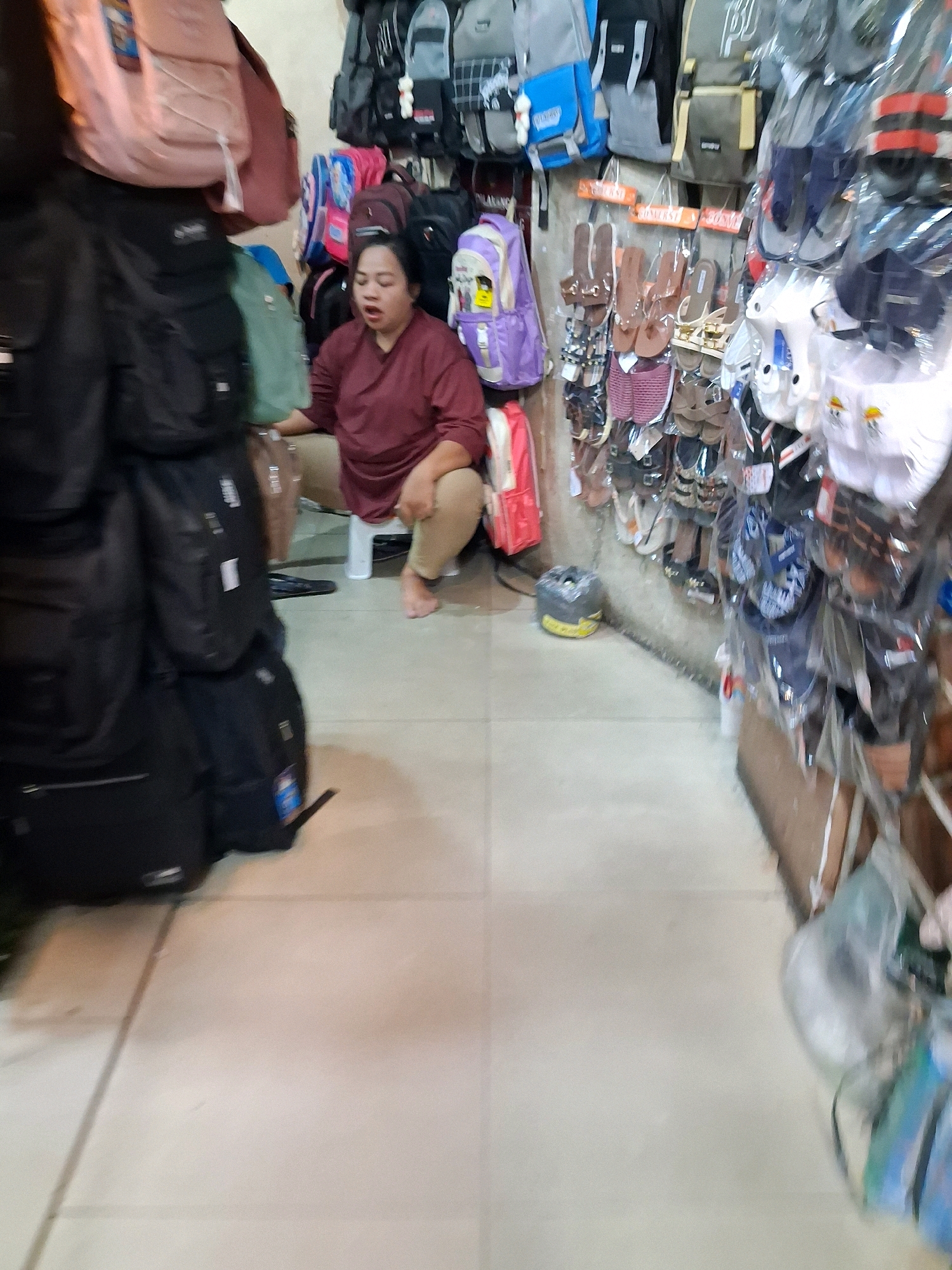 A woman wearing a maroon shirt and tan pants is squatting on a small white stool in a shop. She is looking downwards and appears to be speaking. The shop is filled with merchandise, including numerous backpacks and a display of sandals on the right side of the image. The floor is tiled. This scene is likely from a market stall in Surakarta, Indonesia, given the variety of goods and the informal setup.
